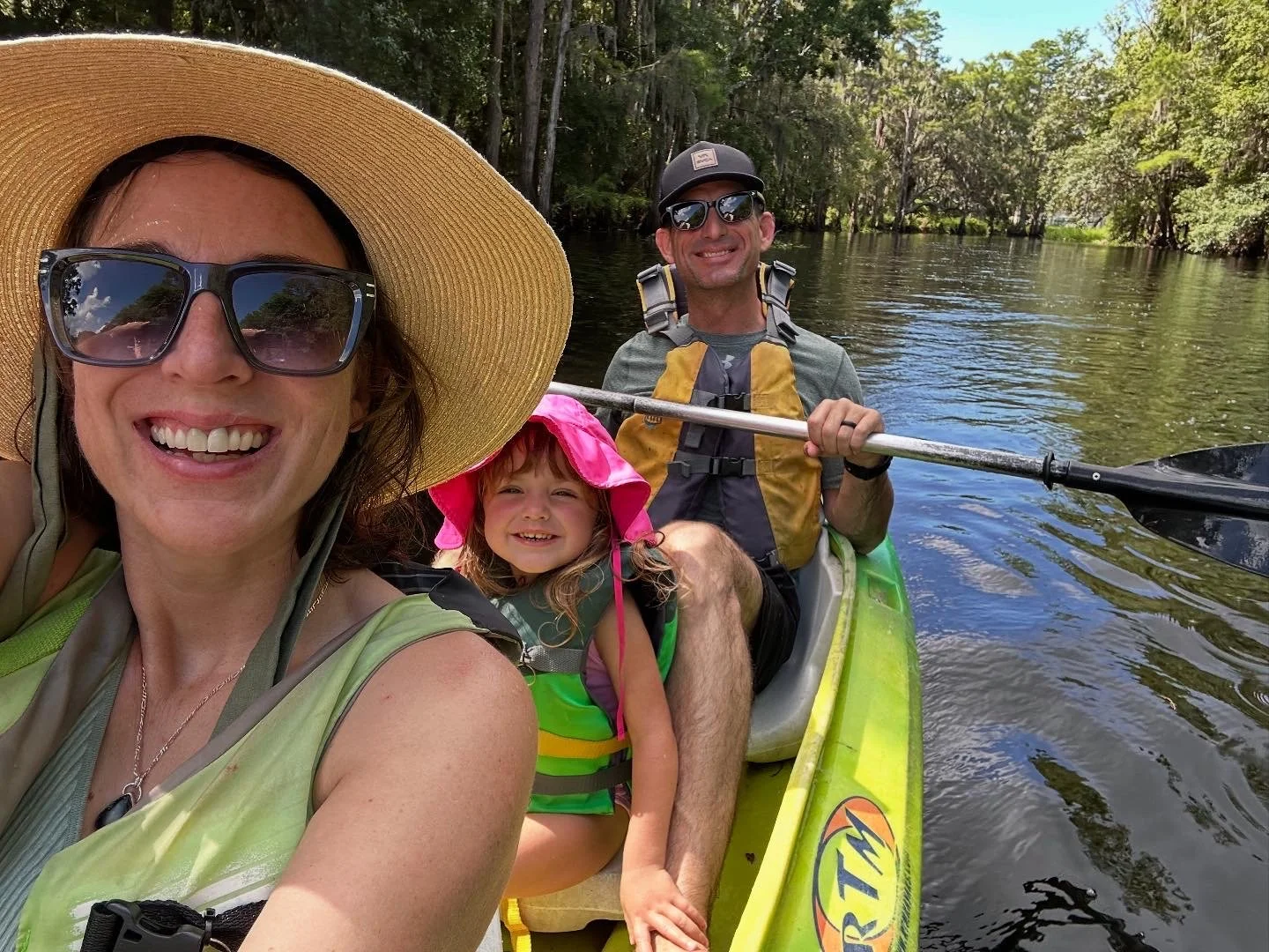 A family of three on a kayaking trip on a river, with trees lining both sides. The woman in the foreground is smiling, wearing sunglasses and a wide-brimmed hat, and taking a selfie. The man behind her is smiling, wearing sunglasses and a cap, holding a paddle. A young girl is sitting between them, wearing a pink hat and life jacket, smiling at the camera.
