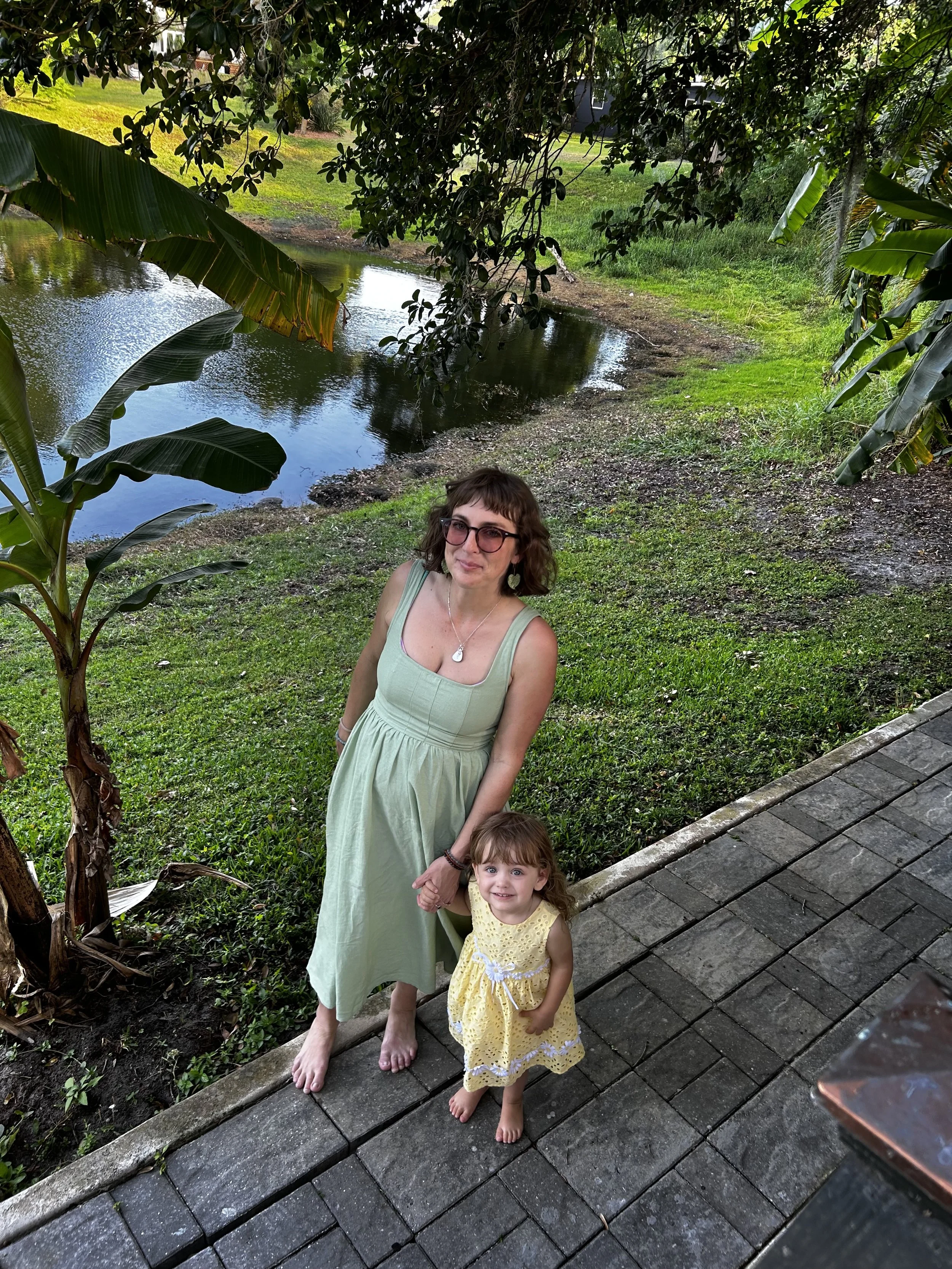 A woman in a light green dress and a young girl in a yellow dress holding hands outdoors by a pond, surrounded by greenery, with trees and a grassy area in the background.