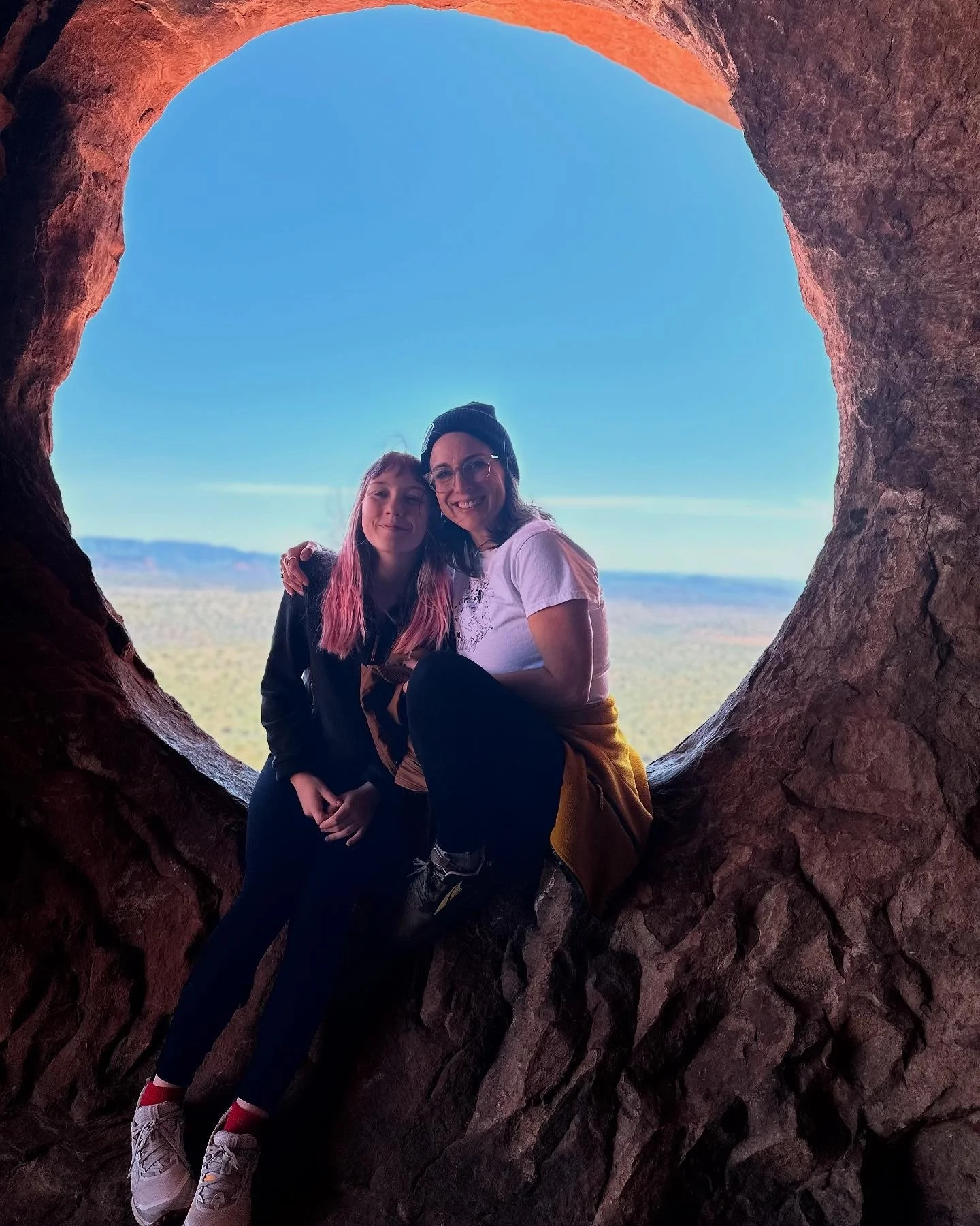 Two women sitting inside a large, heart-shaped rock formation with a scenic view of desert landscape and blue sky in the background.