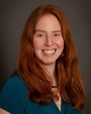 A smiling woman with long red hair wearing a blue top against a gray background.