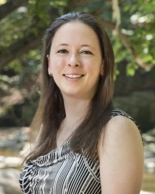 A woman with long brown hair smiling outdoors with trees in the background.