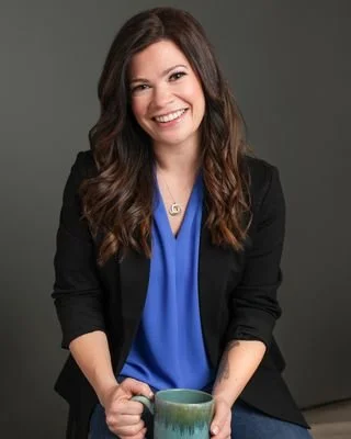 Smiling woman with long brown hair, wearing a black blazer and blue top, sitting and holding a green mug against a gray background.