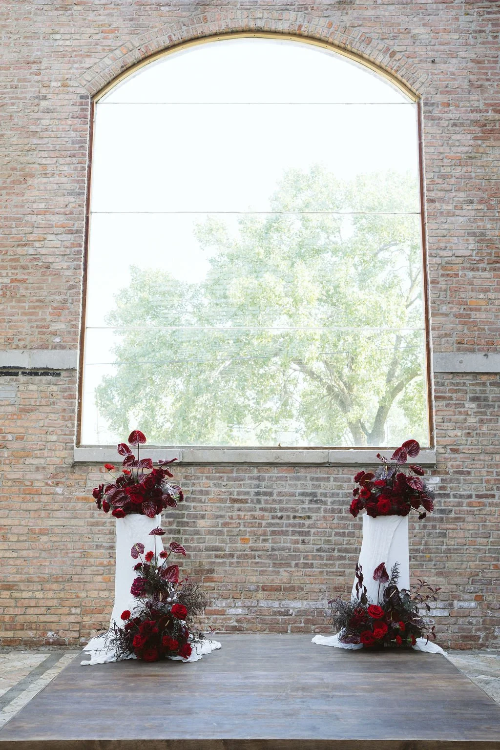 Two floral arrangements on tall stands with red flowers and dark leaves, placed on a wooden platform in front of a large arched window with a brick wall background.