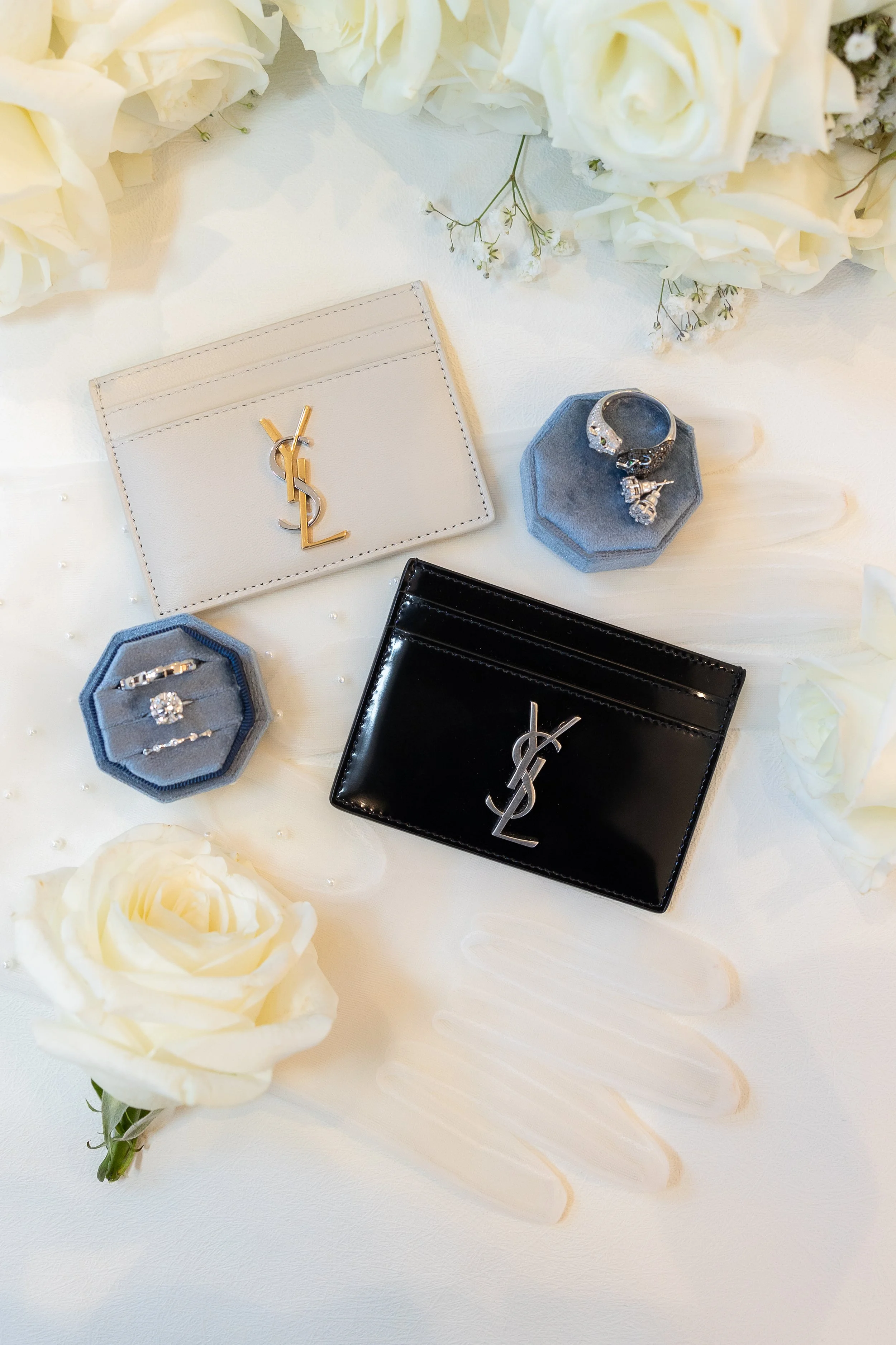 Luxury jewelry and accessories including Yves Saint Laurent and YSL cardholders, rings, and rings in blue velvet holders surrounded by white roses and floral decorations.