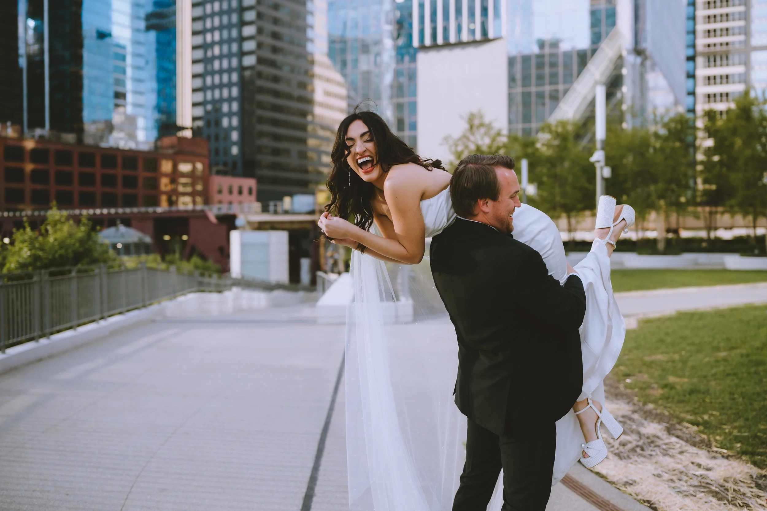 A couple in formal wedding attire outdoors in an urban park, with the man carrying the woman on his back, both smiling and laughing, city skyscrapers in the background.