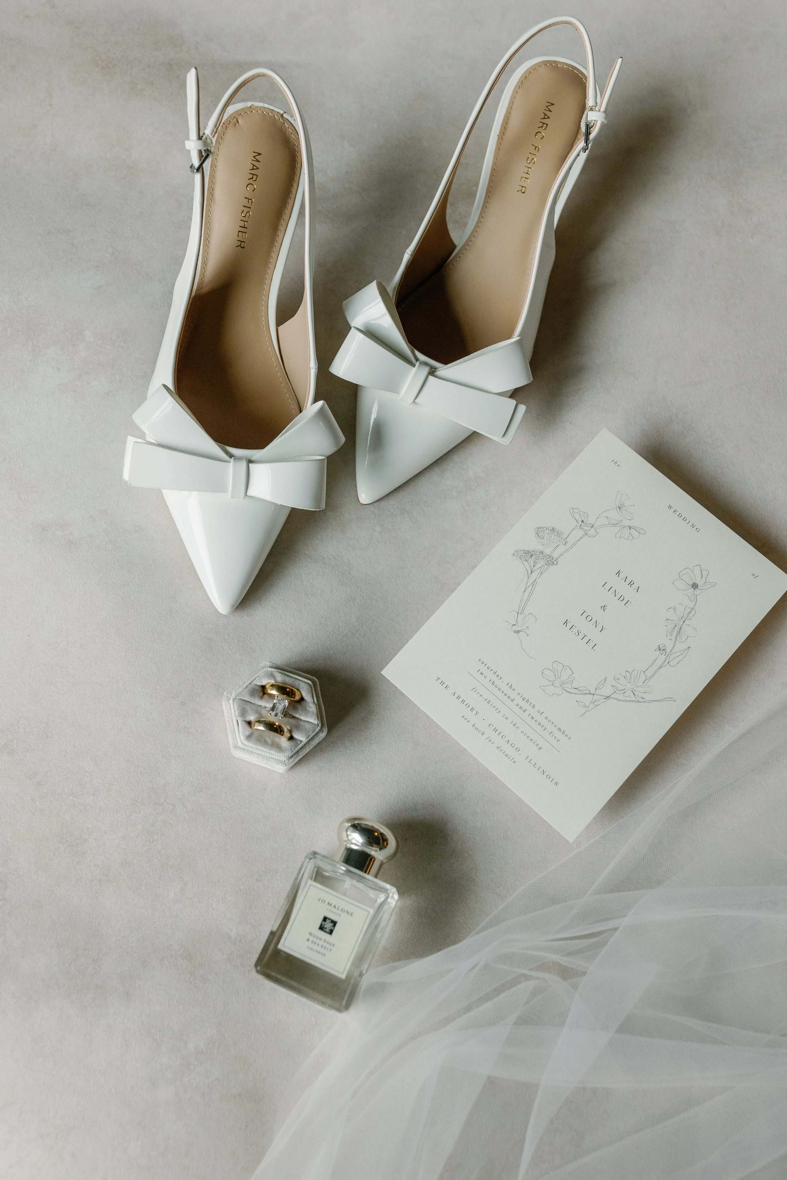 White high-heeled shoes with bows, wedding invitation, rings in a ring box, perfume bottle, and tulle fabric on a light surface.