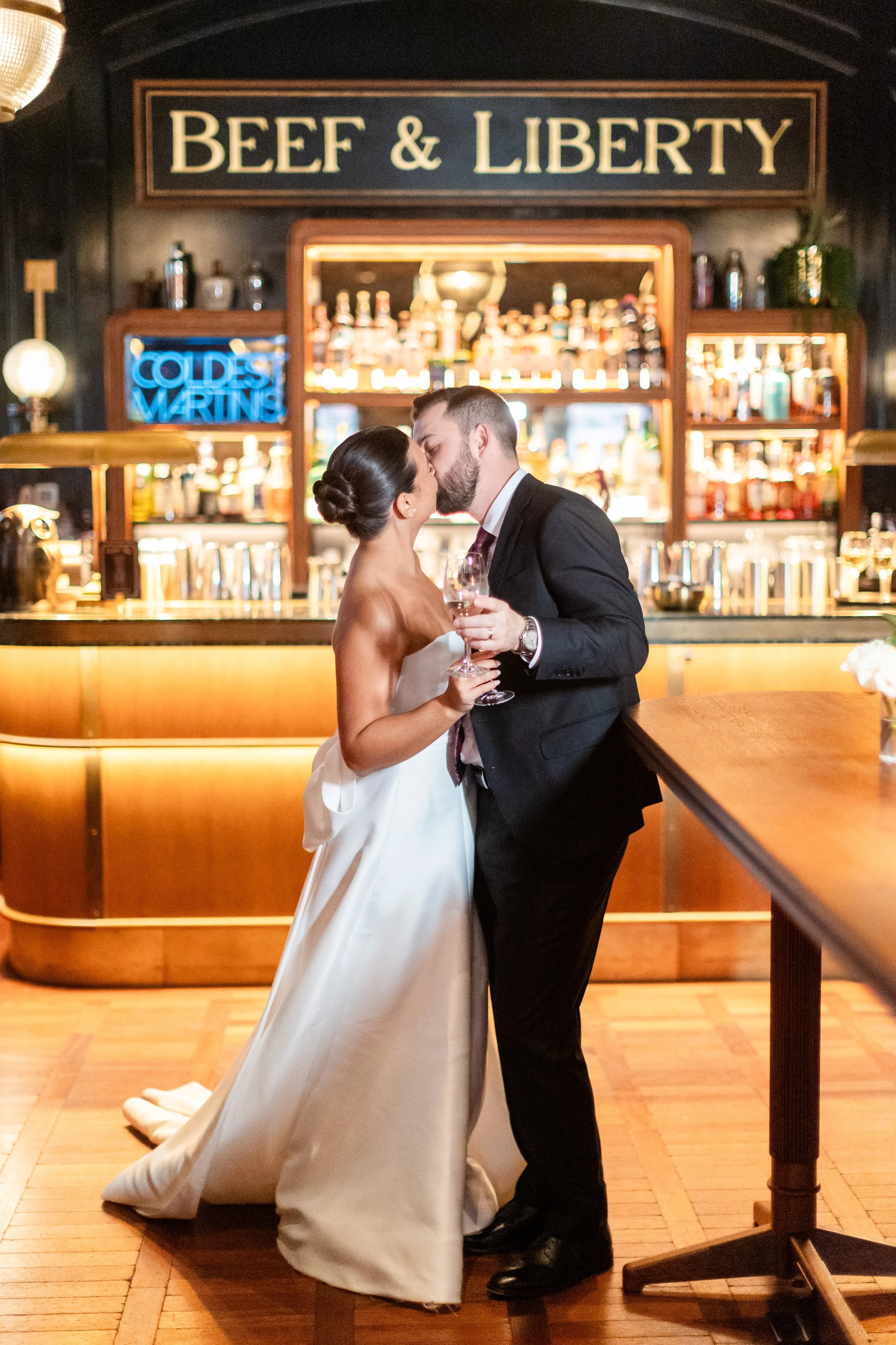 A bride and groom sharing a kiss at a bar, holding glasses of wine, with a sign reading "Beef & Liberty" and a neon sign that says "Coldest Waiting" in the background.
