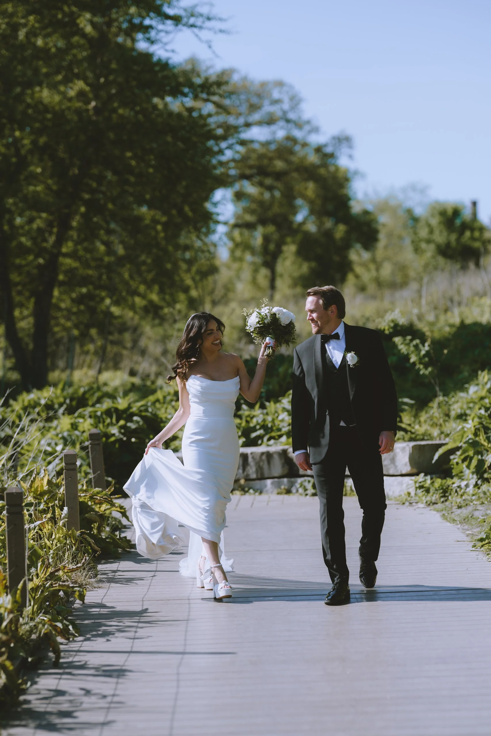 A bride in a white wedding dress and a groom in a black tuxedo walking outdoors on a paved path, with the bride holding a bouquet and smiling at the groom, surrounded by greenery and trees under a blue sky.