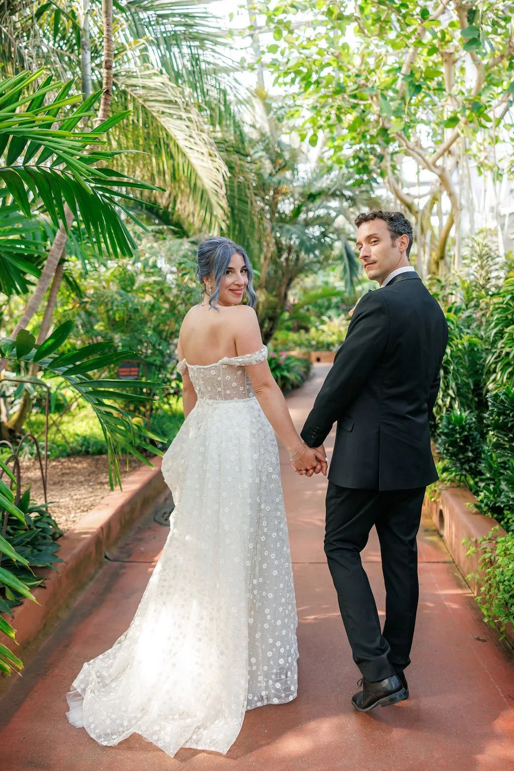 A couple in wedding attire holding hands and looking back, standing in a lush, green tropical garden.