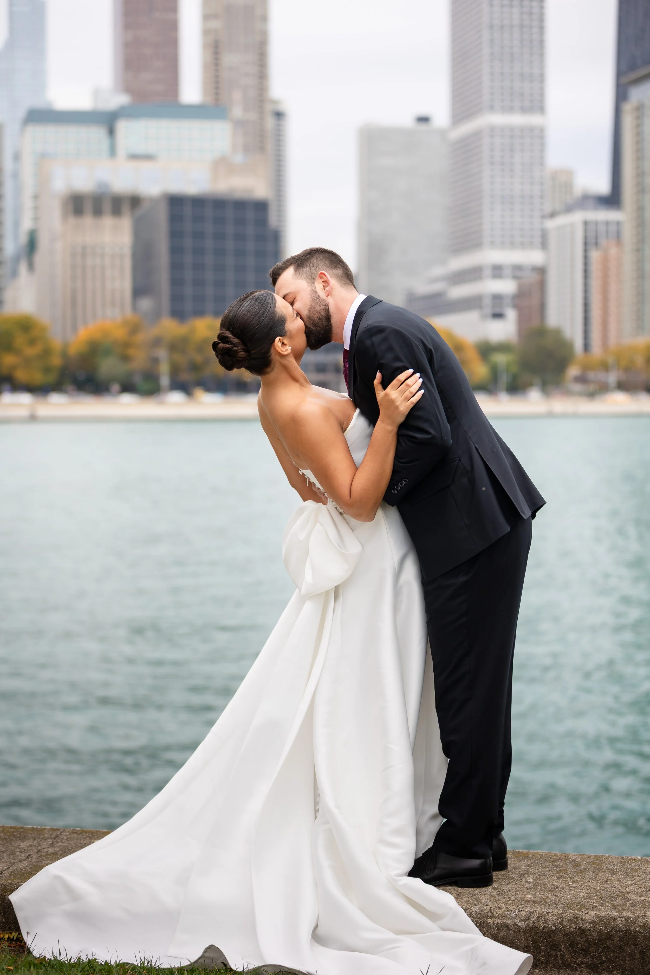 A bride and groom kissing by water with a city skyline in the background.