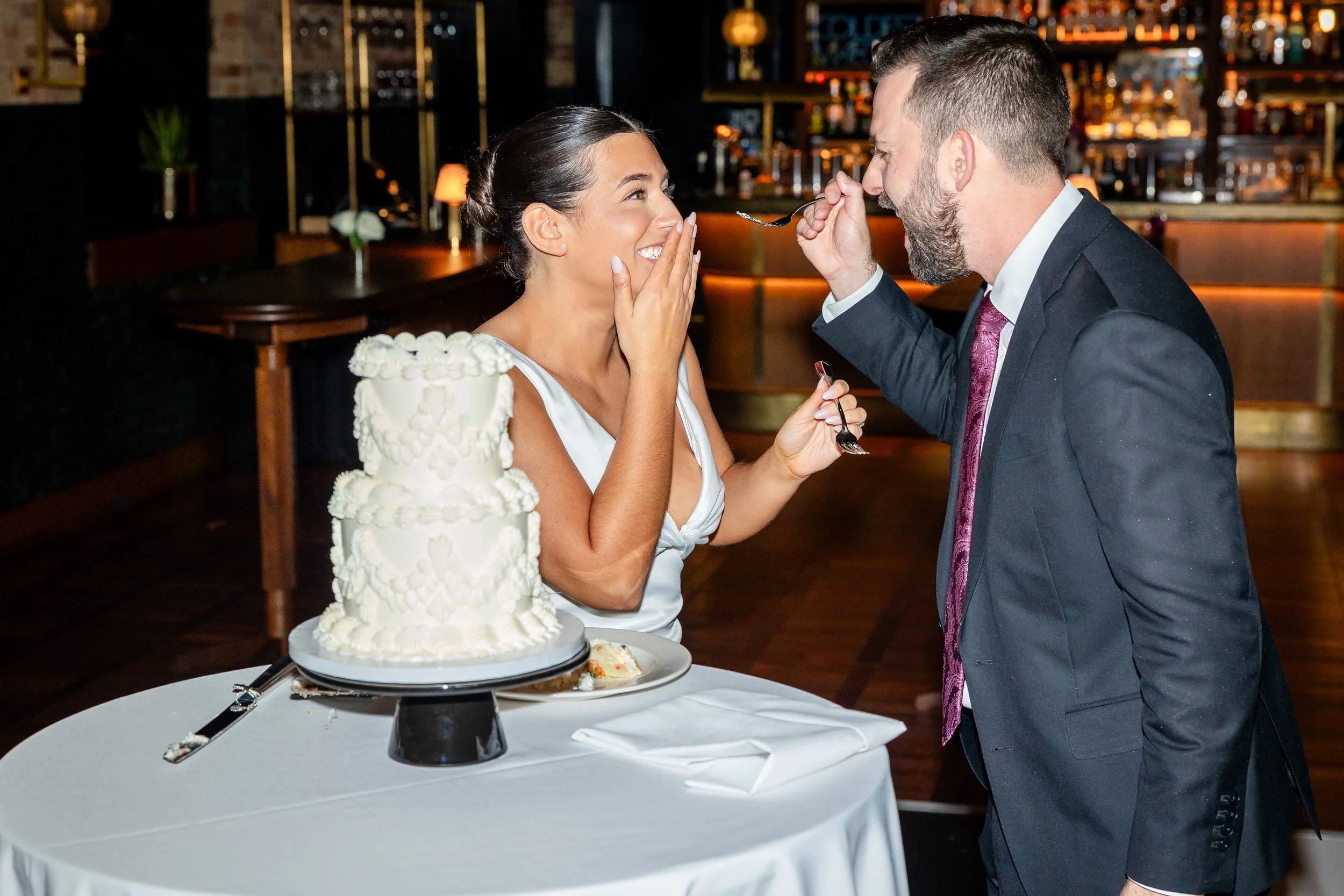 A groom feeding a bride with a spoon at a wedding reception, with a wedding cake on the table.