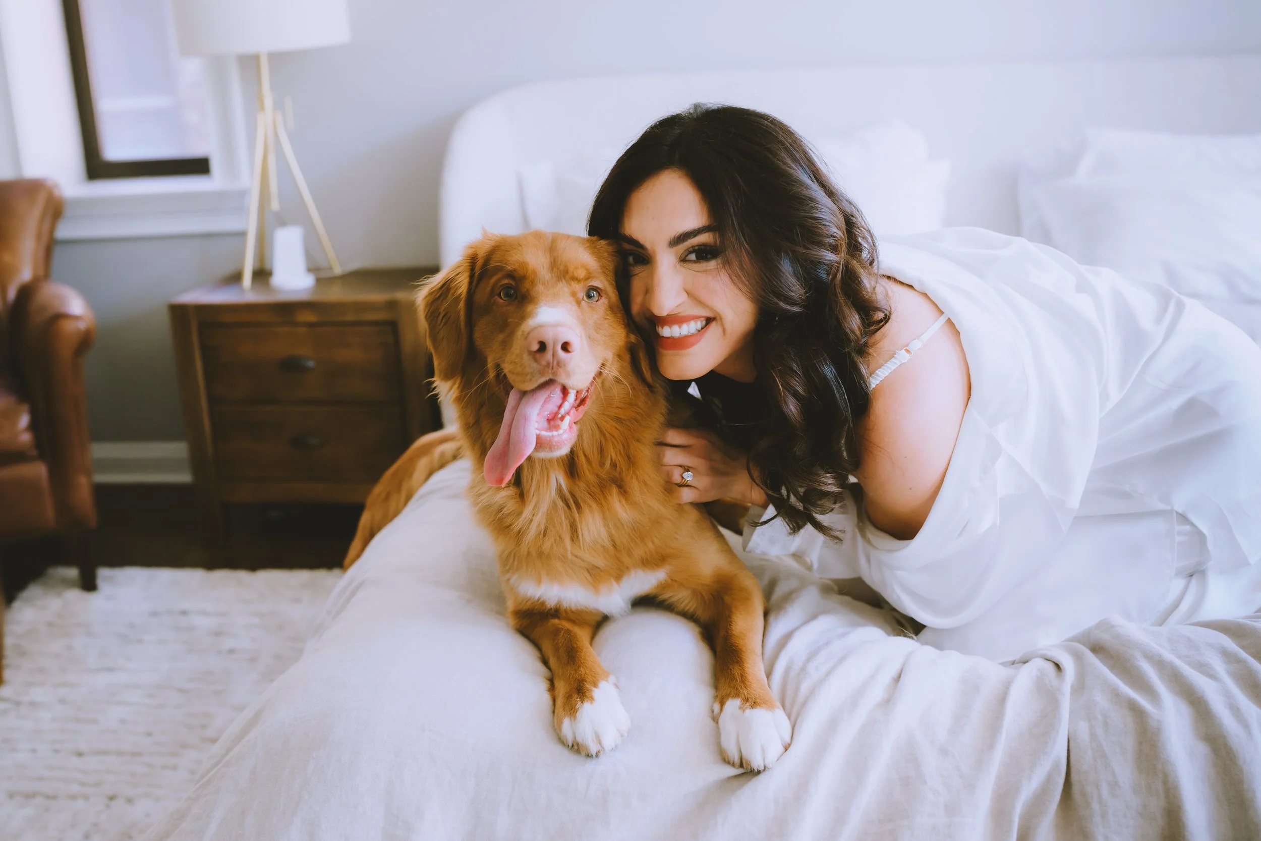 A woman with dark curly hair dressed in white, lying on a bed, smiling, with a happy golden retriever dog sitting beside her in a bright bedroom.