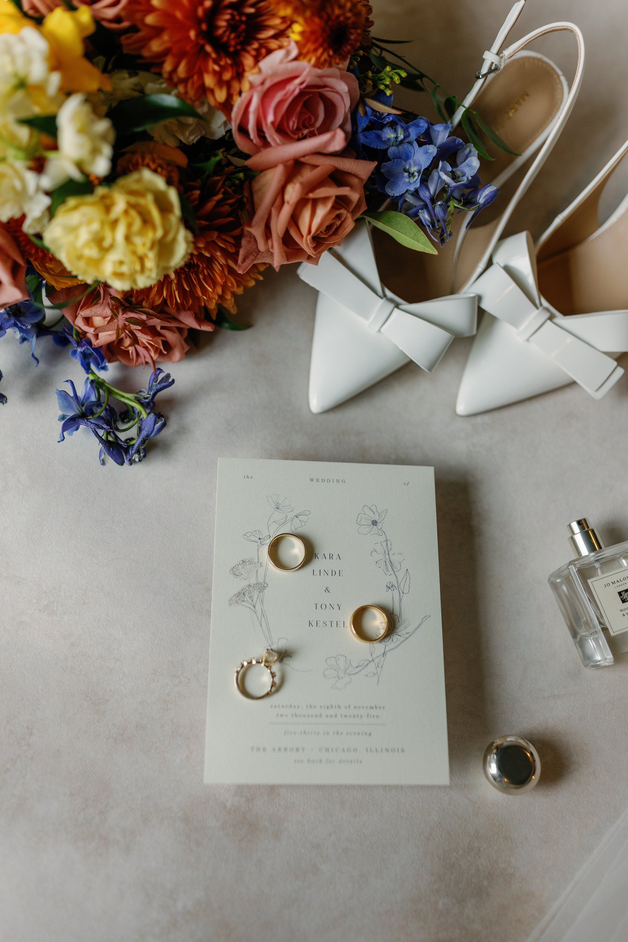 A wedding invitation surrounded by a bouquet of mixed flowers, white high-heeled shoes with bows, perfume bottles, and rings on the invitation.