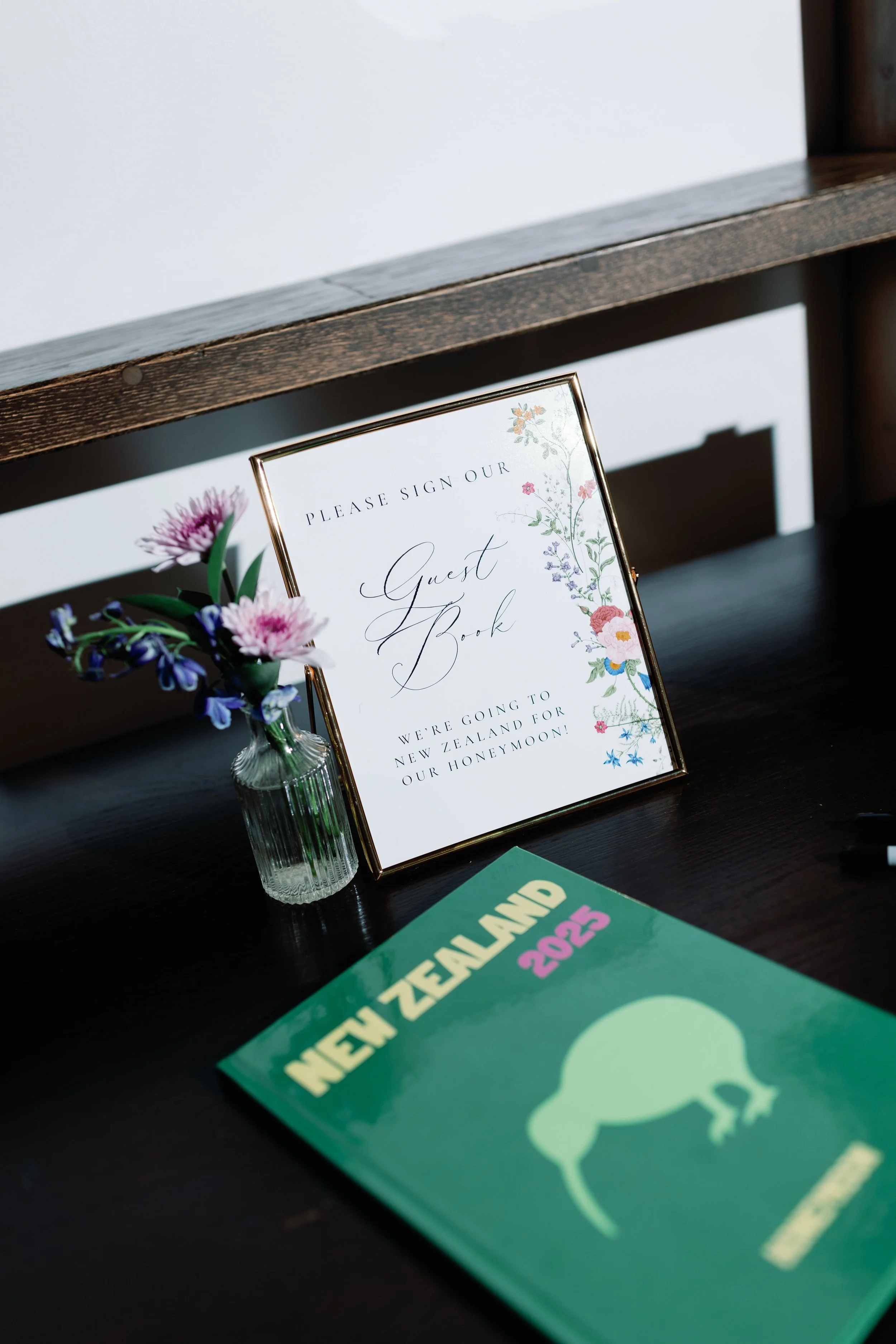 A framed sign that reads 'Please sign our guest book. We're going to New Zealand for our honeymoon,' placed on a dark table. Next to the sign, there is a small glass vase with pink and purple flowers. In the foreground, a green book titled 'New Zeala