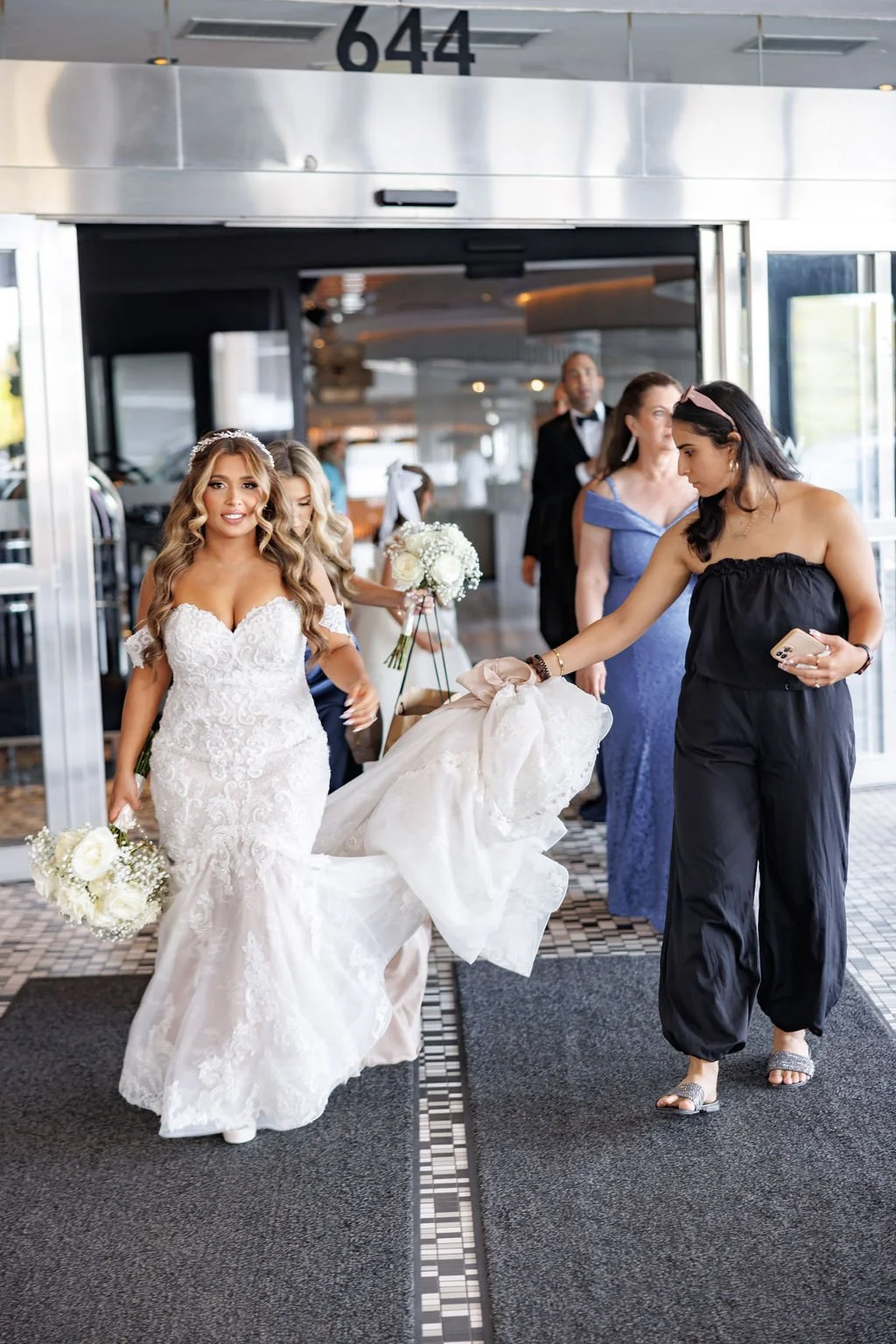 Bride in a white lace wedding dress holding a bouquet walking through the entrance of a building, with bridesmaids and guests in the background.