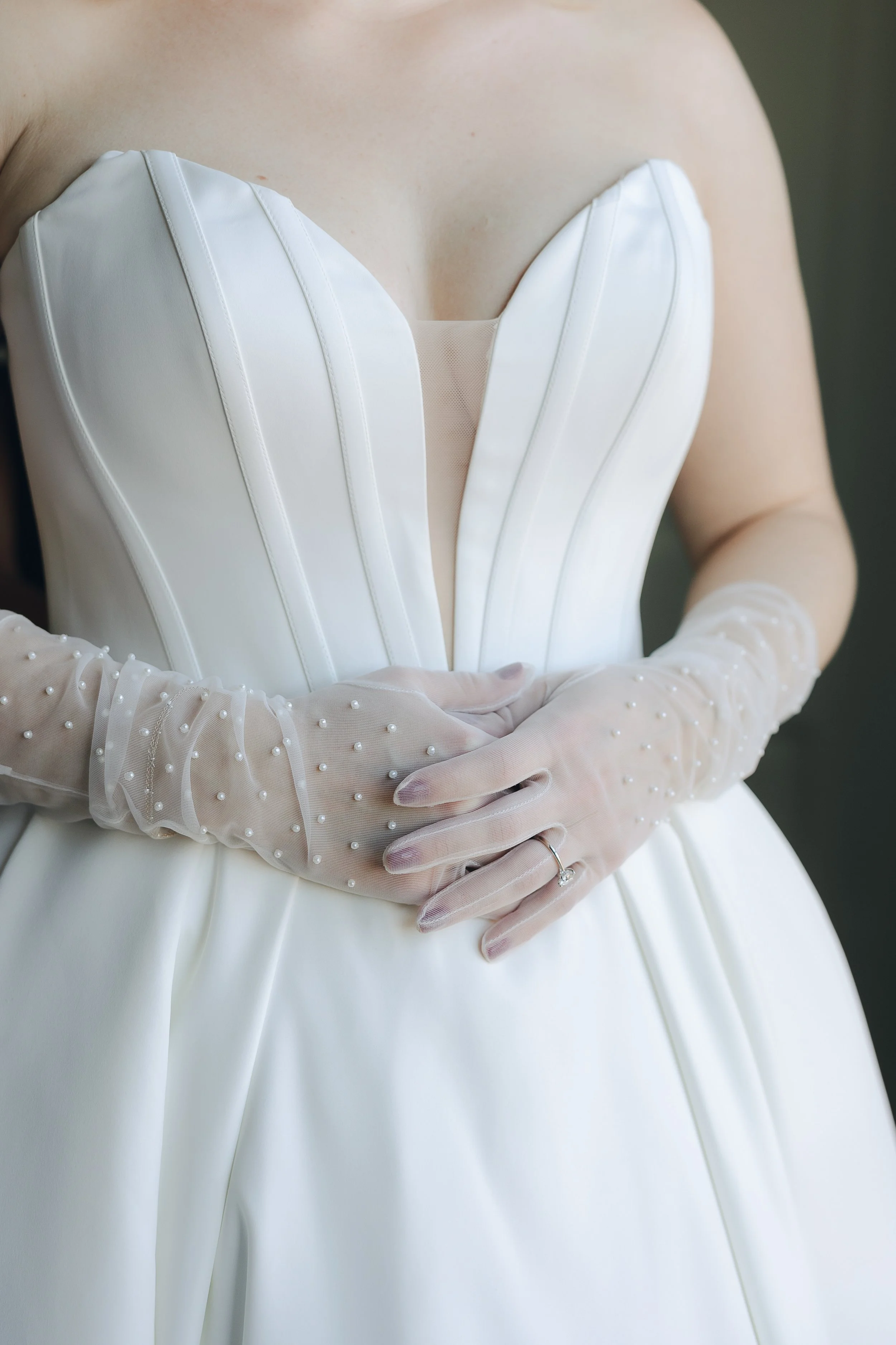 Close-up of a woman in a white wedding dress and sheer gloves with pearl embellishments, holding her hands at her waist.