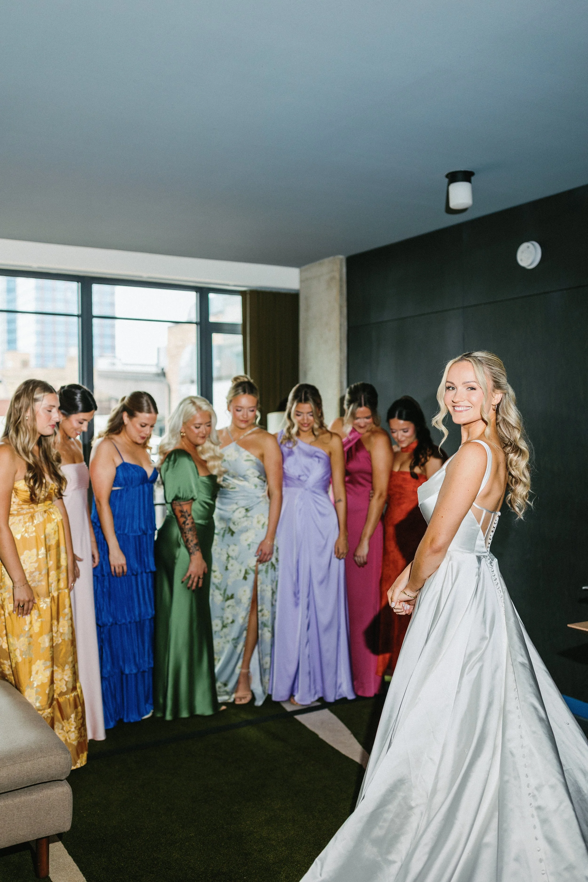 Bride in a white wedding gown with long curly hair smiling at the camera, surrounded by her bridesmaids in colorful dresses standing in a line in a modern room with large windows.