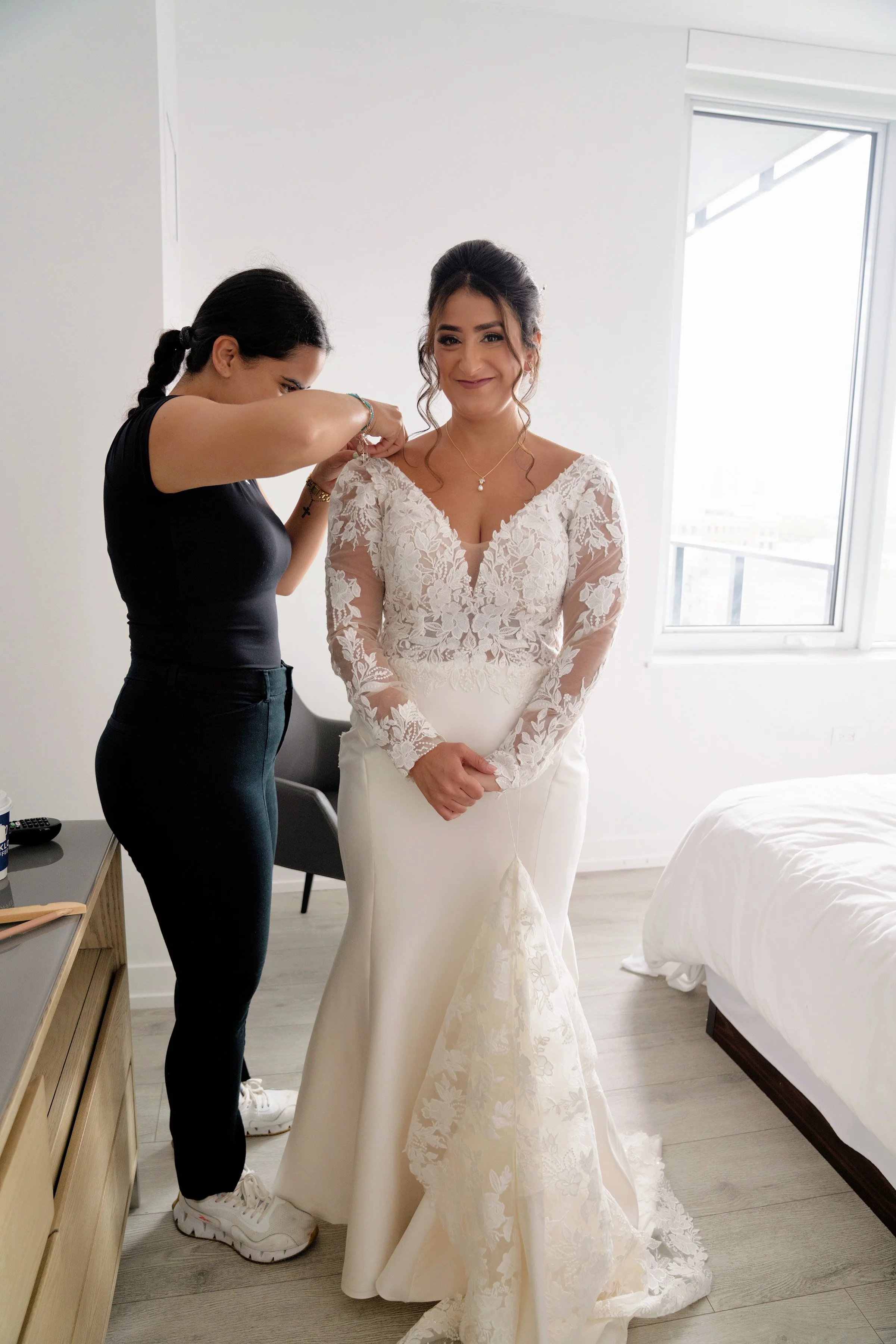 A bride in a white lace wedding dress is smiling while a woman helps her get dressed, adjusting her dress's shoulder strap in a bright room with a window and bed.