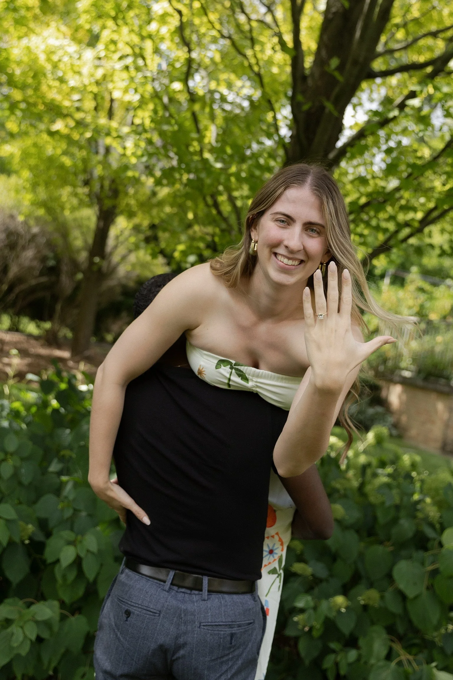A woman showing off her engagement ring outside in a lush green park with trees and bushes.