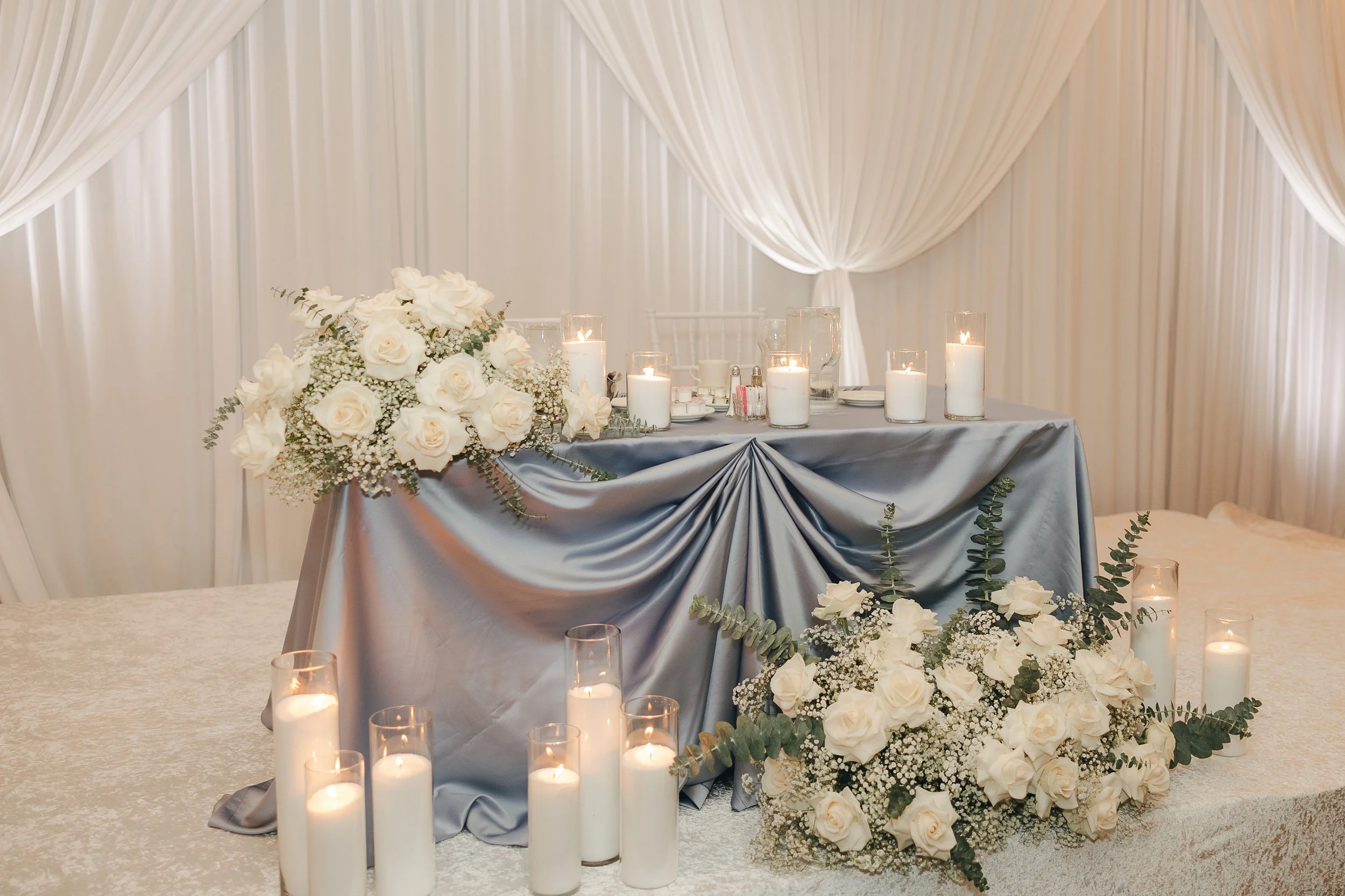 Elegant wedding table with a gray satin tablecloth, floral arrangements of white roses and baby's breath, surrounded by candles in glass holders, set against cream-colored drapes.