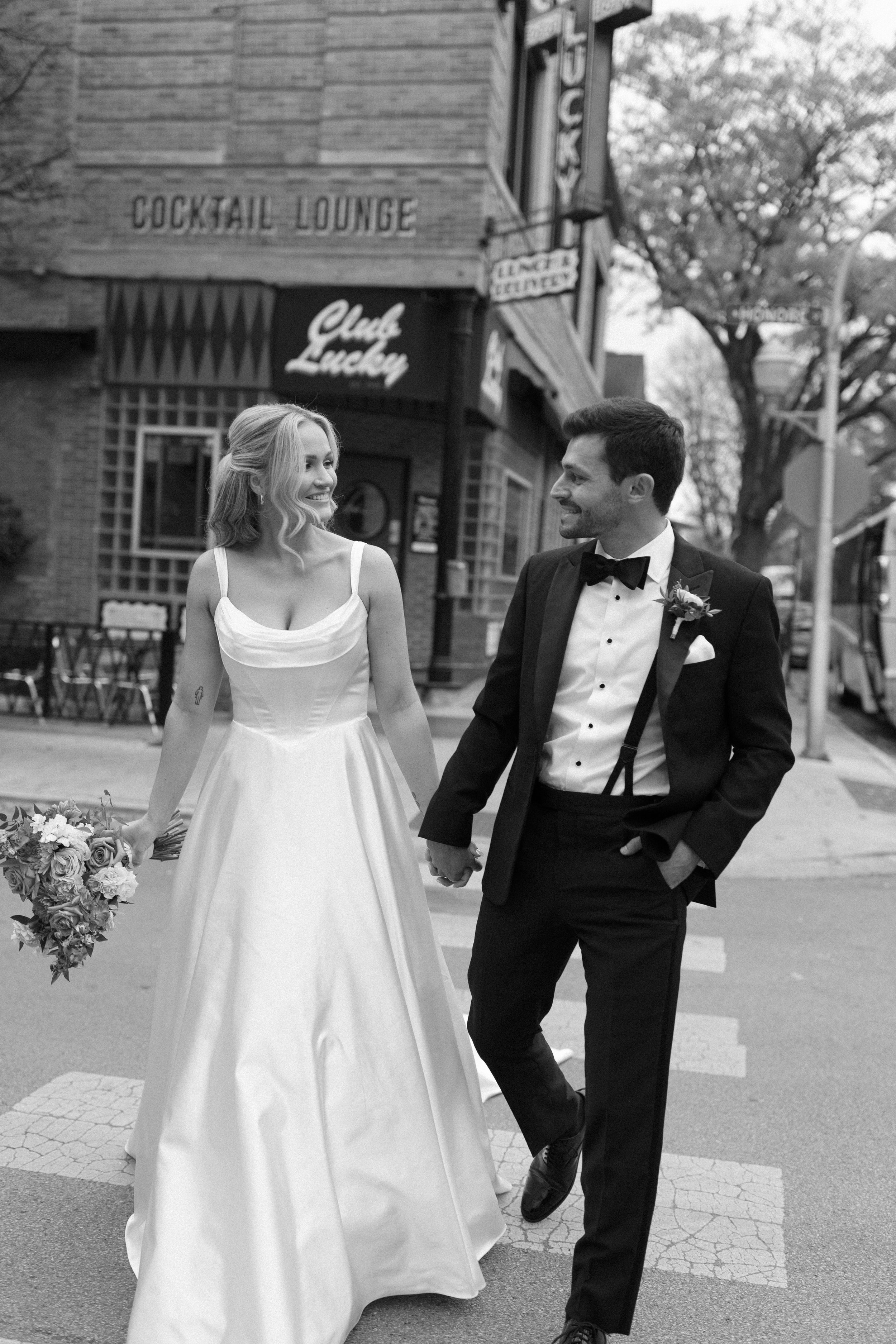 A black-and-white photo of a bride and groom walking hand in hand on a city street, smiling at each other. The bride is wearing a white wedding gown and holding a bouquet of flowers, while the groom is dressed in a tuxedo with a bow tie and boutonnie
