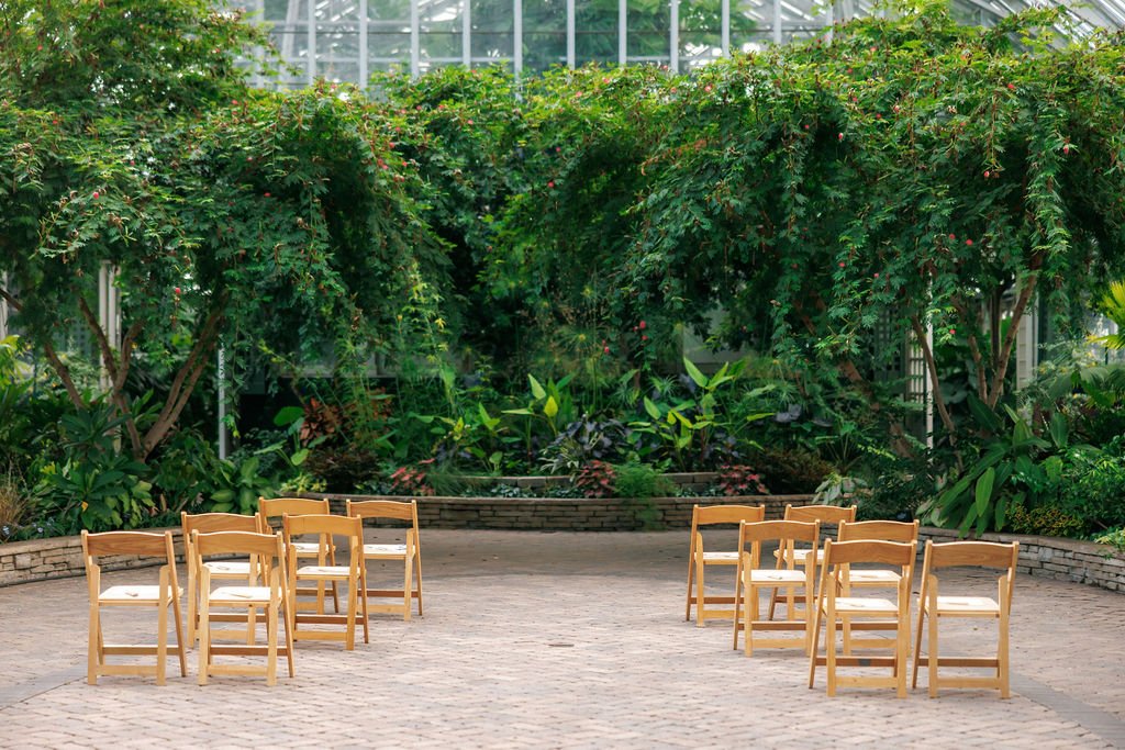 Rows of wooden chairs arranged outdoors in a garden setting with lush greenery and trees in the background.