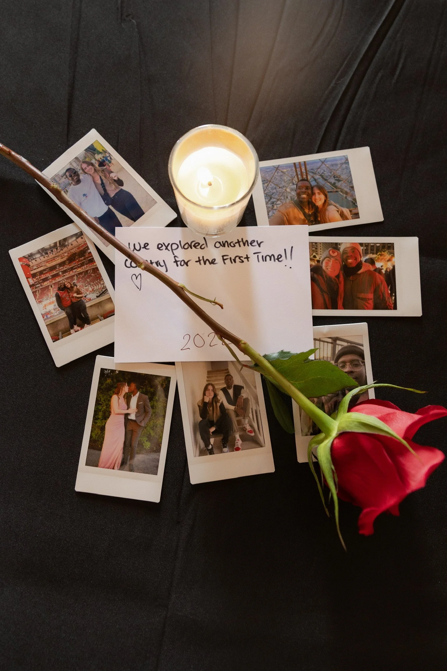A red rose, a lit candle, and several printed photographs of people are arranged on a black surface. A handwritten note that says, "We explored another country for the first time!!" and the year 2023 is also present.