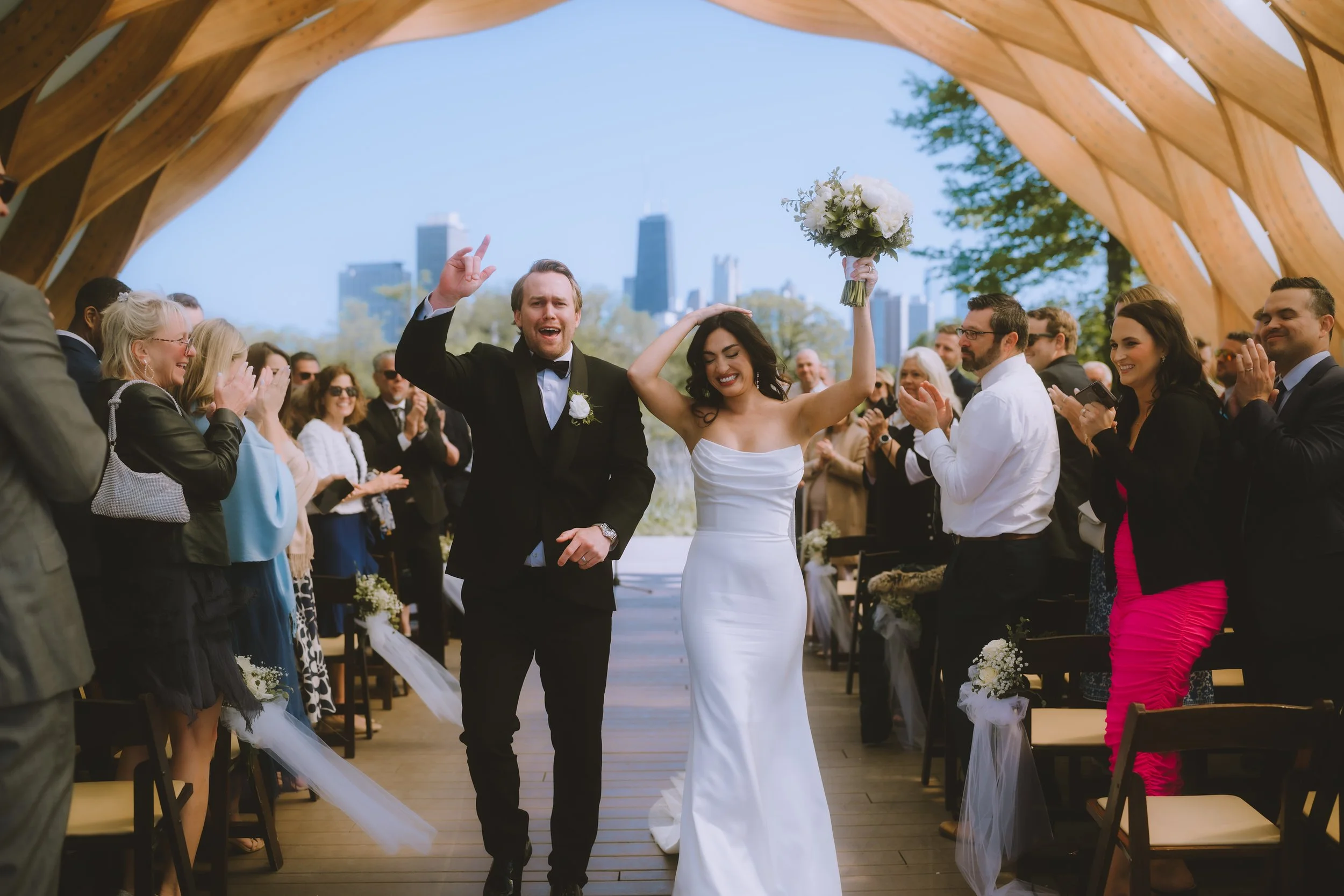 A bride and groom celebrating after their wedding ceremony outdoors, with guests clapping and cheering around them.