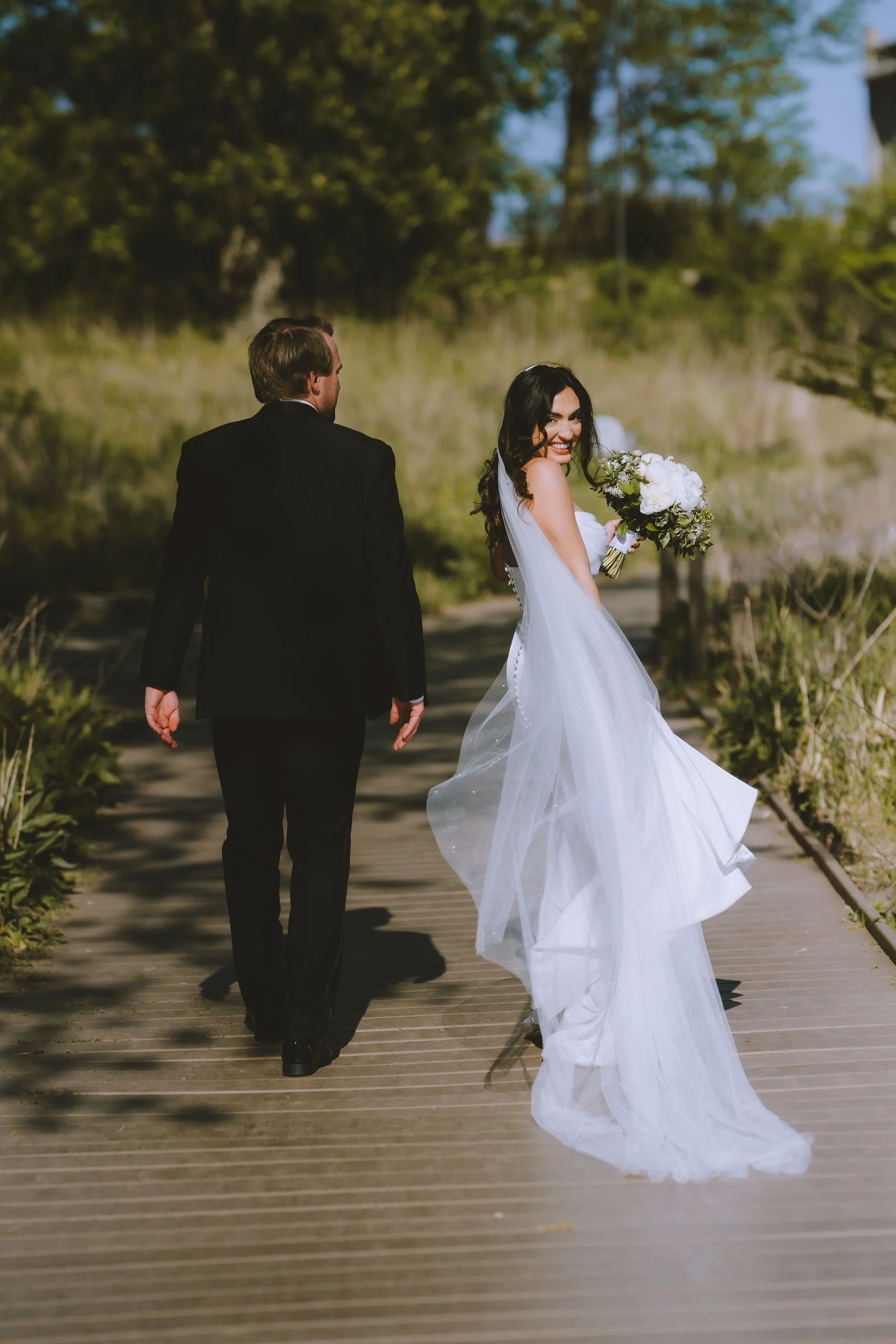 A bride in a white wedding dress holding a bouquet, smiling and looking back, walking on a wooden pathway beside a groom in a black suit, with greenery and trees in the background.