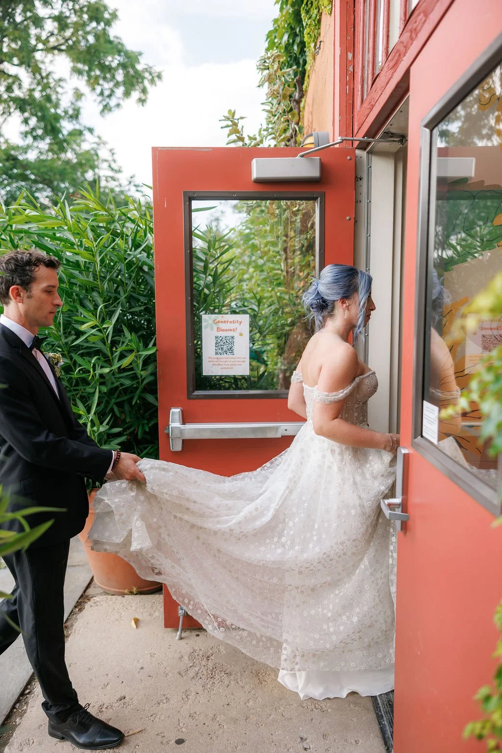 A bride with blue hair in a wedding dress holding the door open for a groom in a tuxedo who is pulling her train outside a building with green foliage around.
