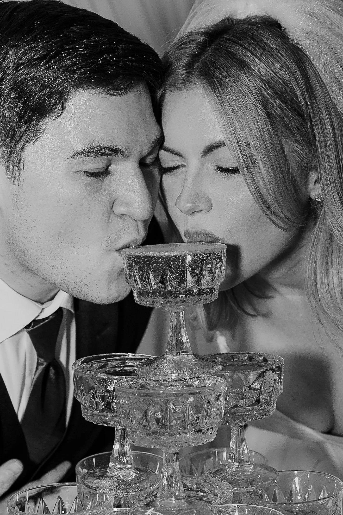 A black and white photo of a bride and groom, both with their eyes closed, about to clink glasses on a tiered glass serving stand, with the bride wearing a veil.