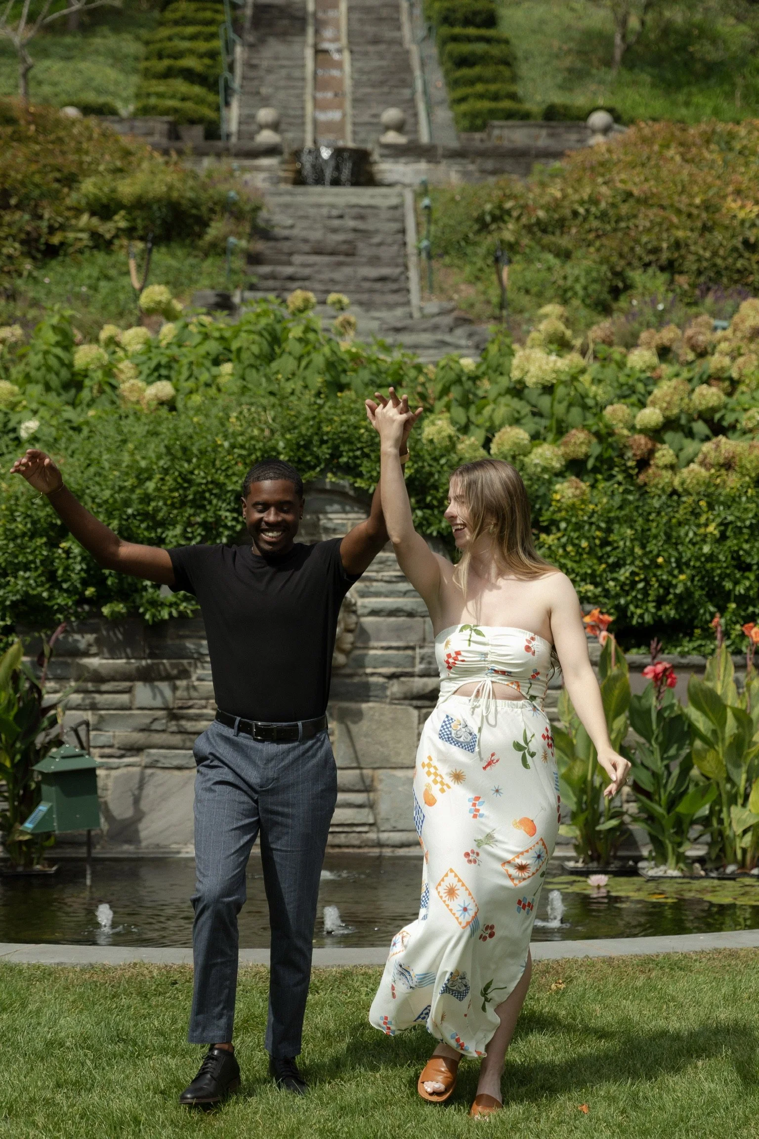 A man and woman dancing and smiling in a garden with green plants, flowers, a fountain, a stone staircase, and a landscaped hillside in the background.