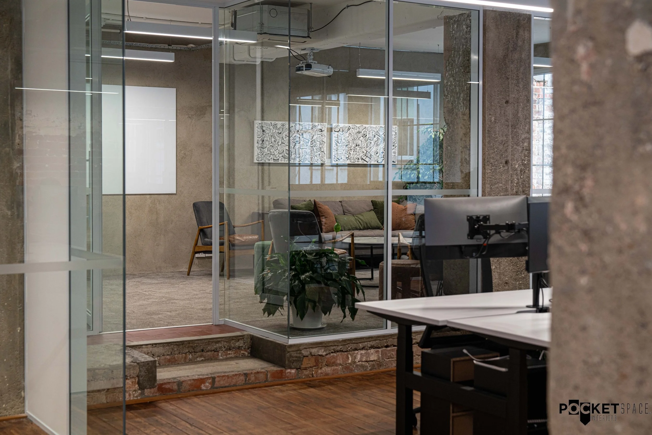 Modern office interior with glass walls, concrete columns, and a potted plant. There is a visible workspace with a computer monitor, and a meeting area with chairs and artwork on the wall. Natural light enters through large windows, and the floor is