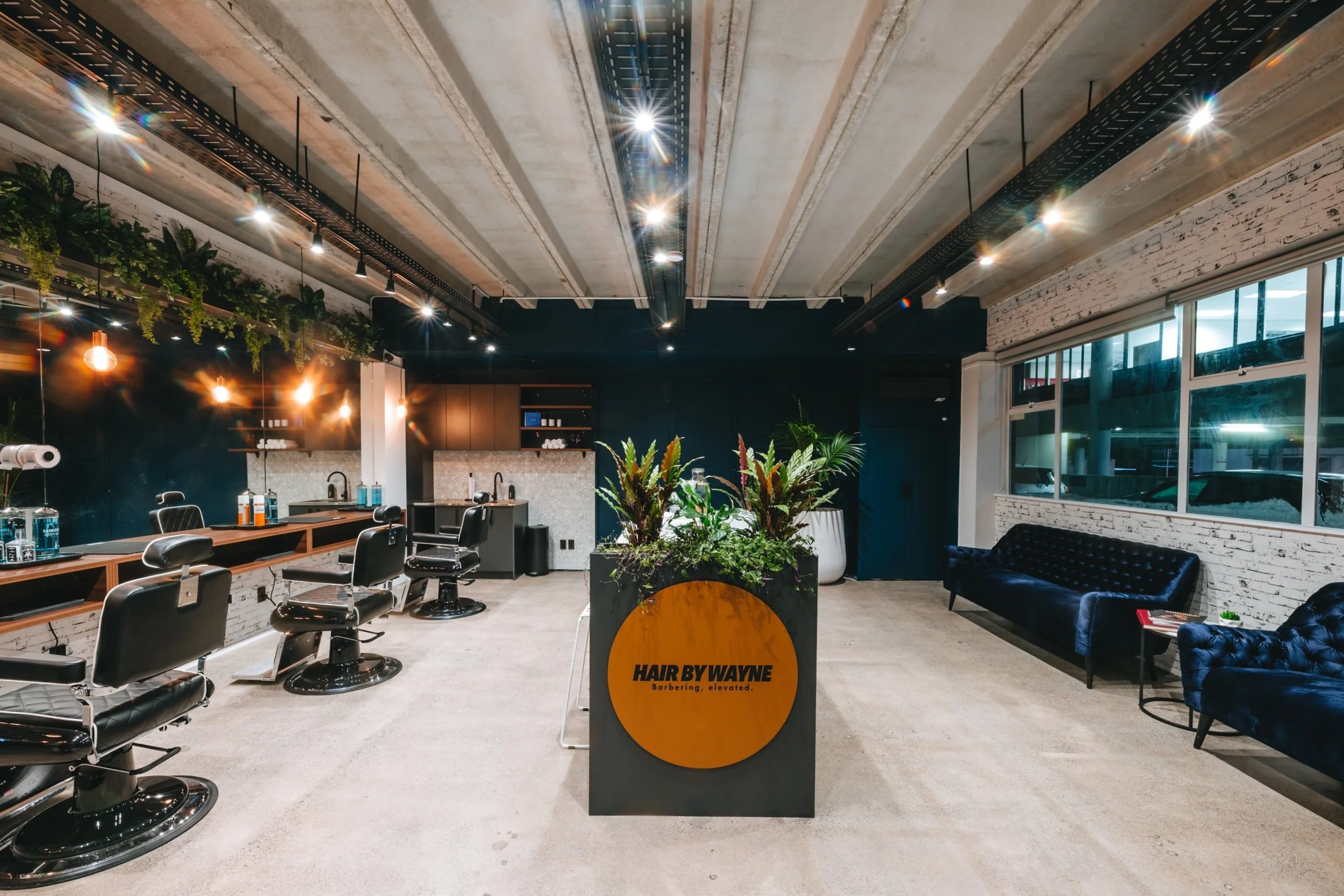 Modern barbershop interior with stylish leather chairs, wooden mirrors, indoor plants, and a reception desk labeled 'HAIR BY WAYNE'. Industrial design with exposed beams and ambient lighting.