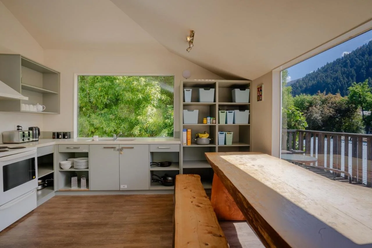 Modern kitchen interior with white cabinets, large window, wooden floor, and open shelves holding kitchen appliances and dishes. A wooden table and bench are positioned near the window, offering a view of lush greenery and mountains.