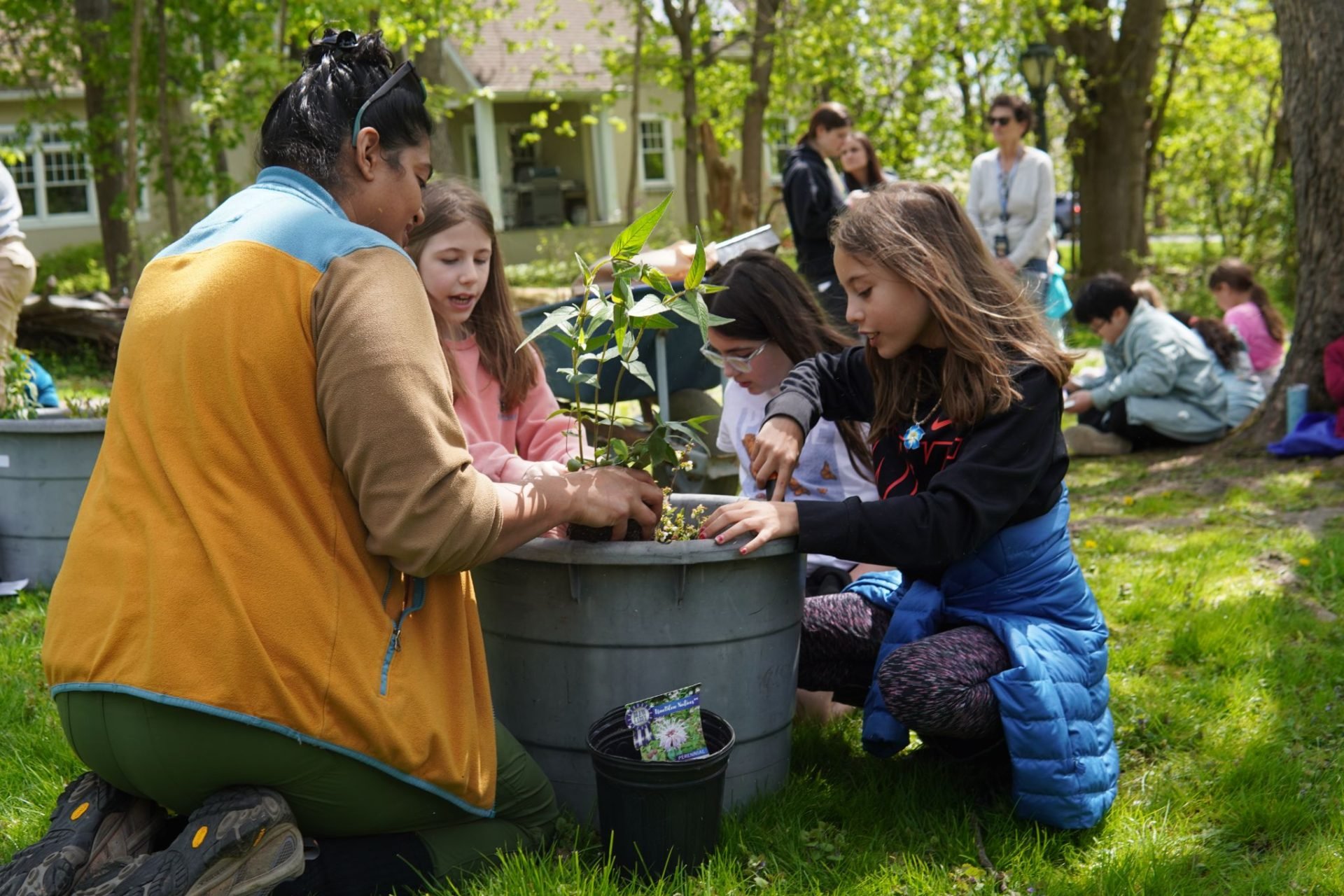 Group of children and adult planting and arranging small plants in a large container outside on grass, with additional people in the background near trees.