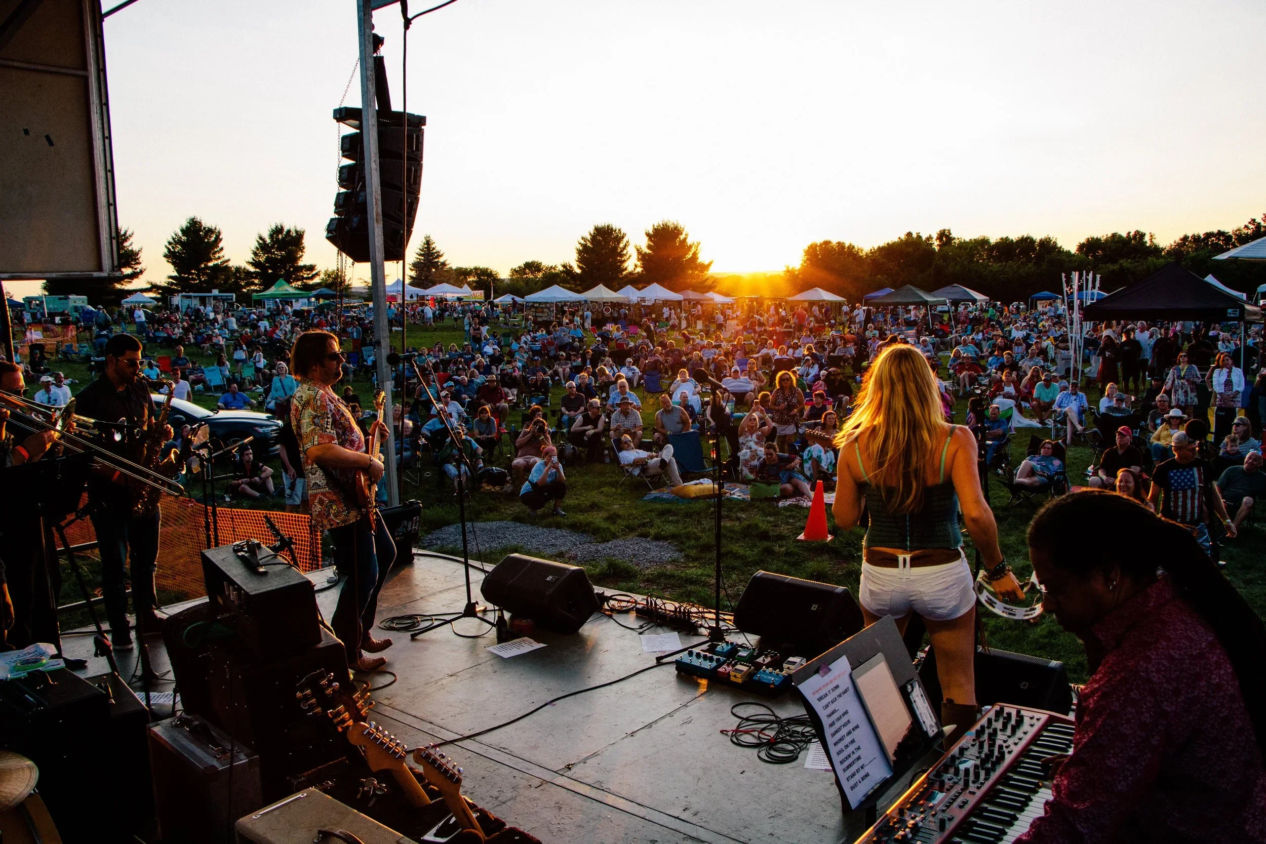 Live outdoor concert at sunset with a band performing on stage and a large audience sitting on grass in front of tents and trees.