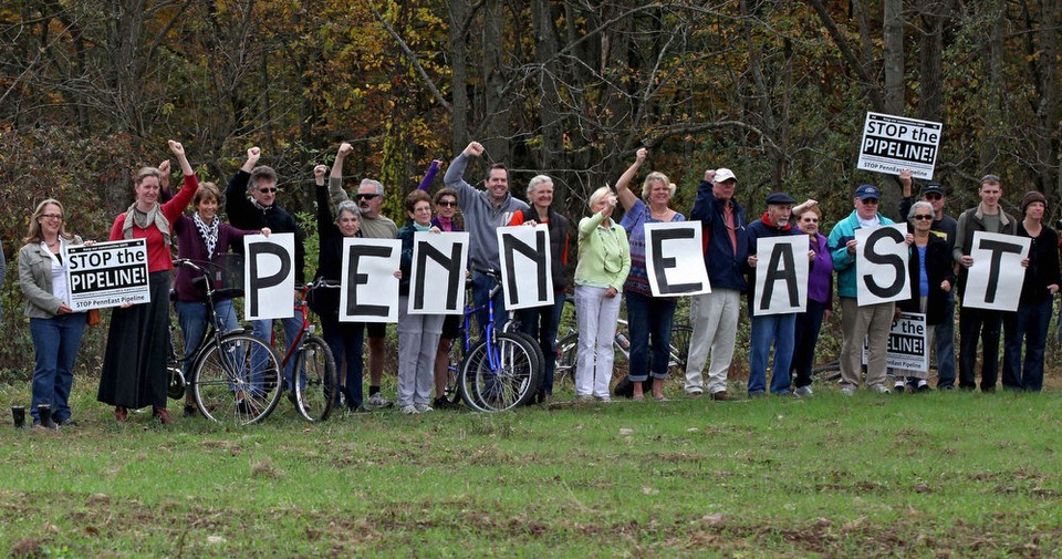 Group of protesters holding signs that spell out "PENNEAST" and signs that say "STOP the PIPELINE!" during a rally in an outdoor setting with trees in the background.