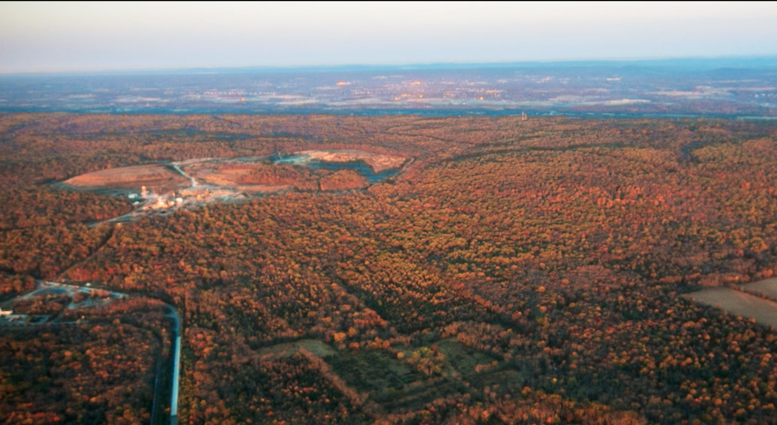 Aerial view of autumnal forest with trees in fall colors, a small lake, and some industrial structures in the distance. Open farmland and distant cityscape on the horizon.