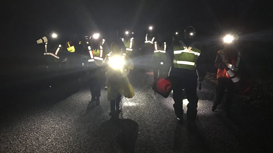 Nighttime scene of emergency responders with reflective vests and flashlights standing on a dark road.