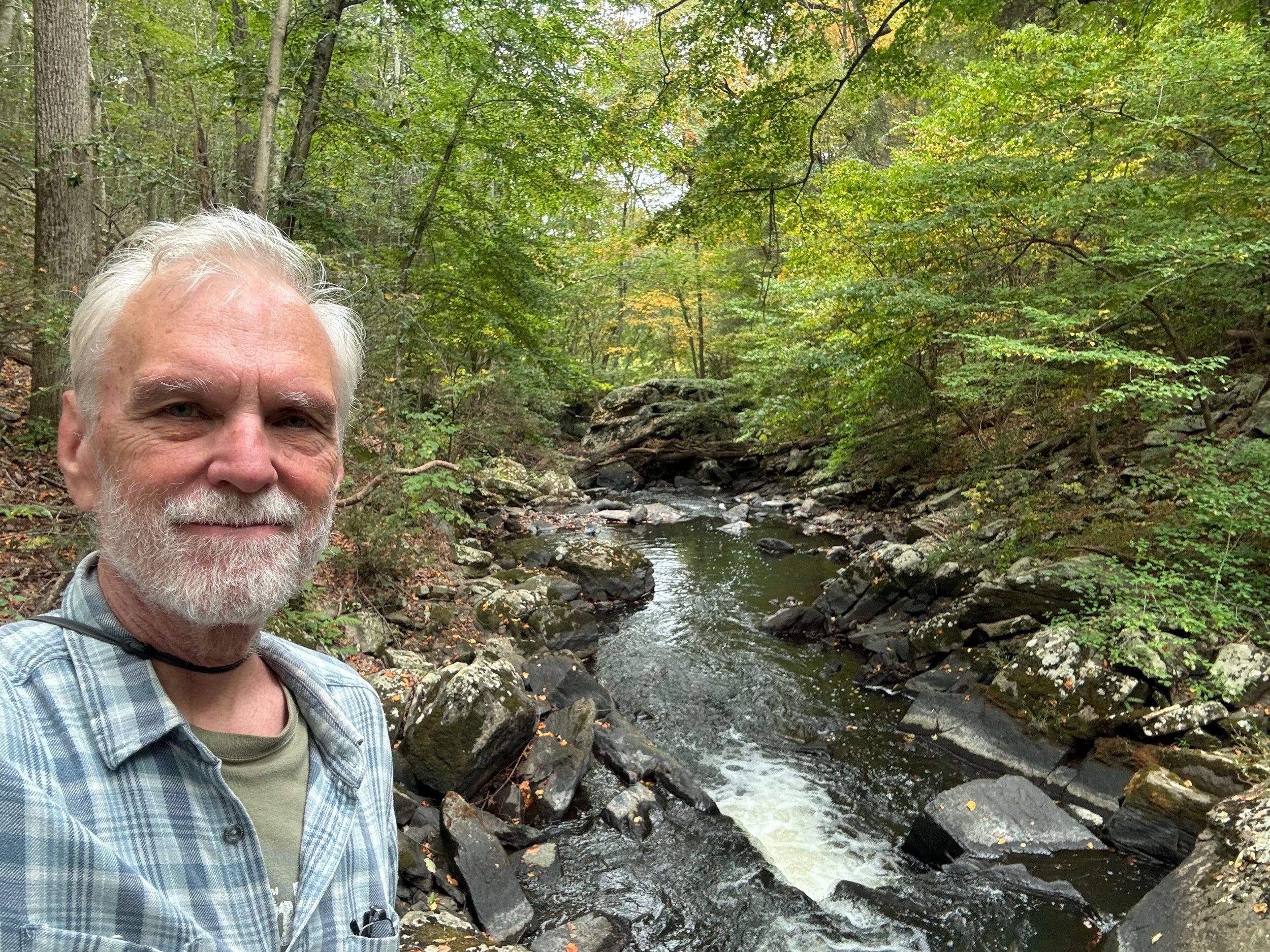 An older man with white hair and beard smiling in front of a rocky creek in a lush, green forest.