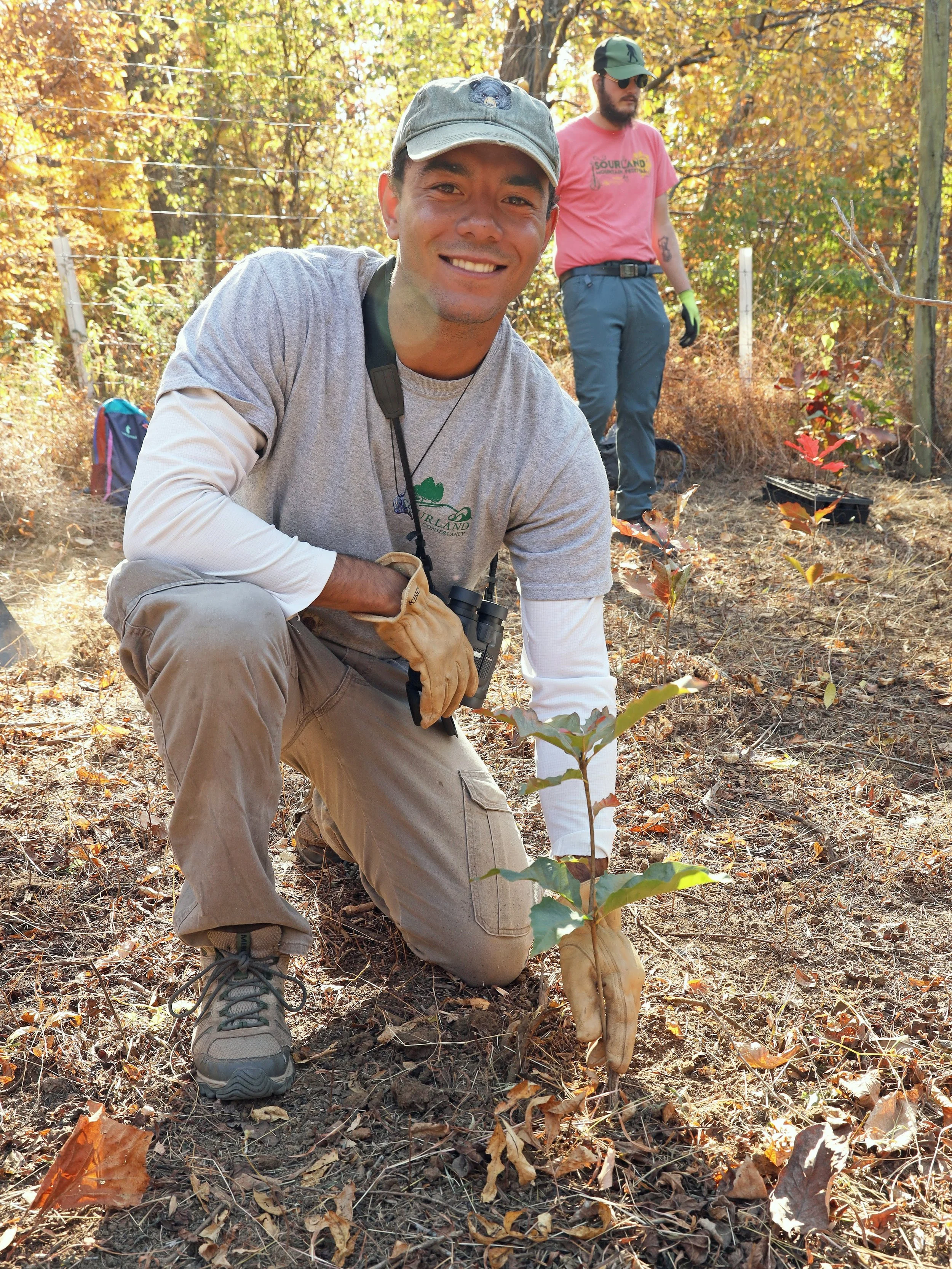 A man kneeling in a wooded area, smiling, with a small tree sapling in front of him. He is wearing a gray cap, gray t-shirt, beige pants, and gloves. Two other people are visible in the background, one wearing a pink shirt and sunglasses, the other partly obscured.