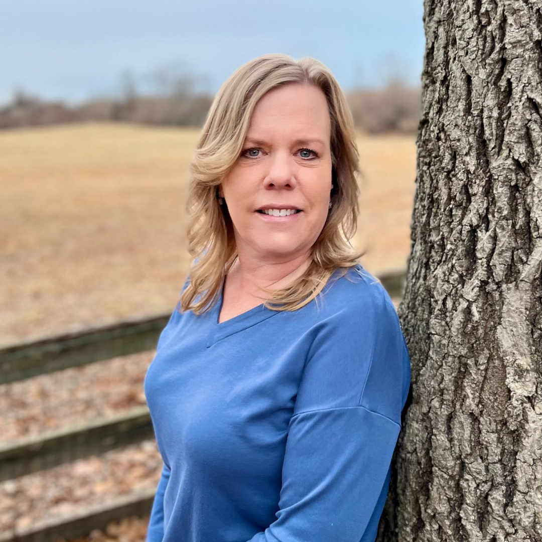 A woman with blonde hair wearing a blue top, leaning against a tree outdoors with a blurred background of a field and a fence.