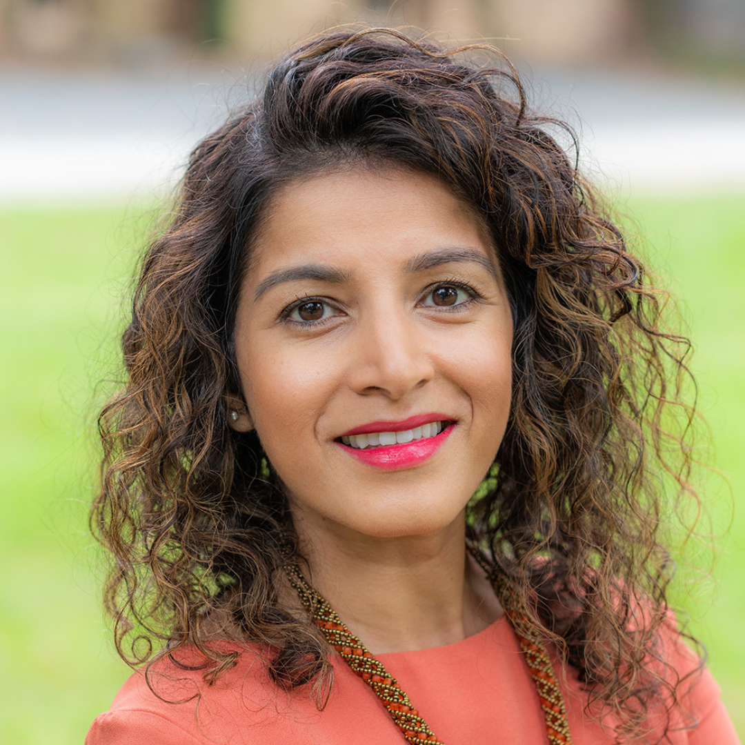 Close-up portrait of a woman with curly brown hair, wearing a coral top, lipstick, and earrings, outdoors with a blurred green background.
