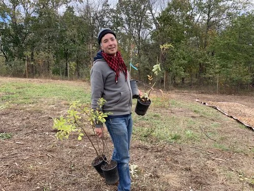 A young man standing outdoors in a cleared yard holding two small potted trees, one in each hand, with trees and a fence in the background.