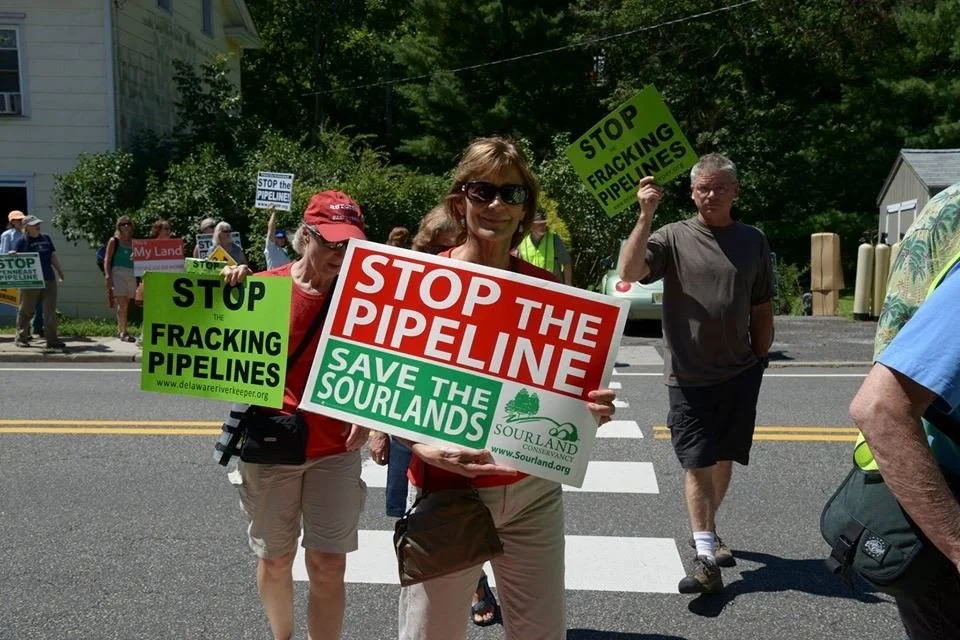 A group of people protesting against fracking pipelines, holding signs that say 'Stop the Pipeline,' 'Stop Fracking Pipelines,' and 'Save the Sourlands' on a street.