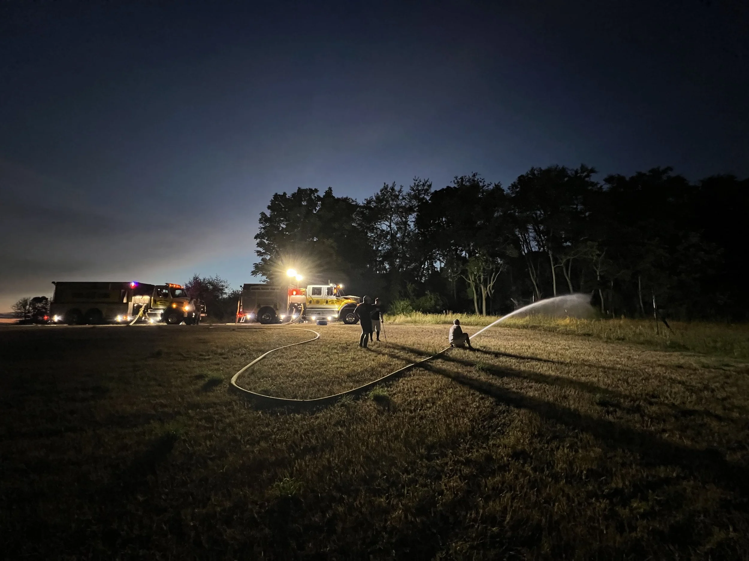 Nighttime scene with firefighters and fire trucks, one person using a hose to spray water on the ground near trees in a field, illuminated by artificial lights.