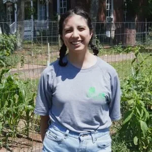 Young woman standing in a garden with green plants and trees in the background, smiling, wearing a gray T-shirt and jeans.