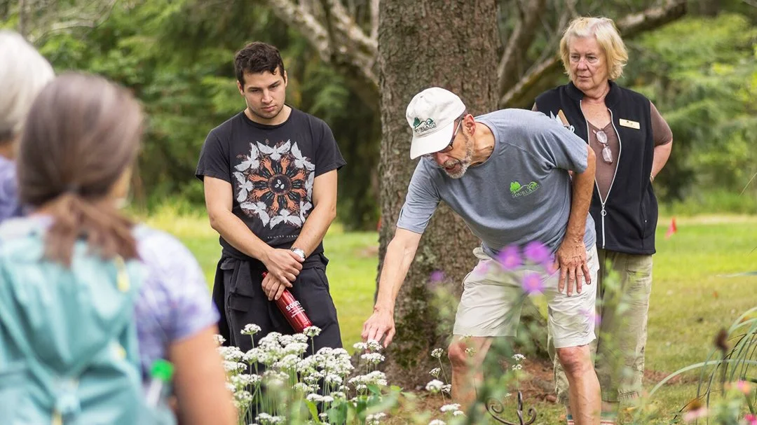 A man in a gray shirt and white cap showing a group of children and an older woman around a garden bed with white flowers, outdoors in a park or garden setting.