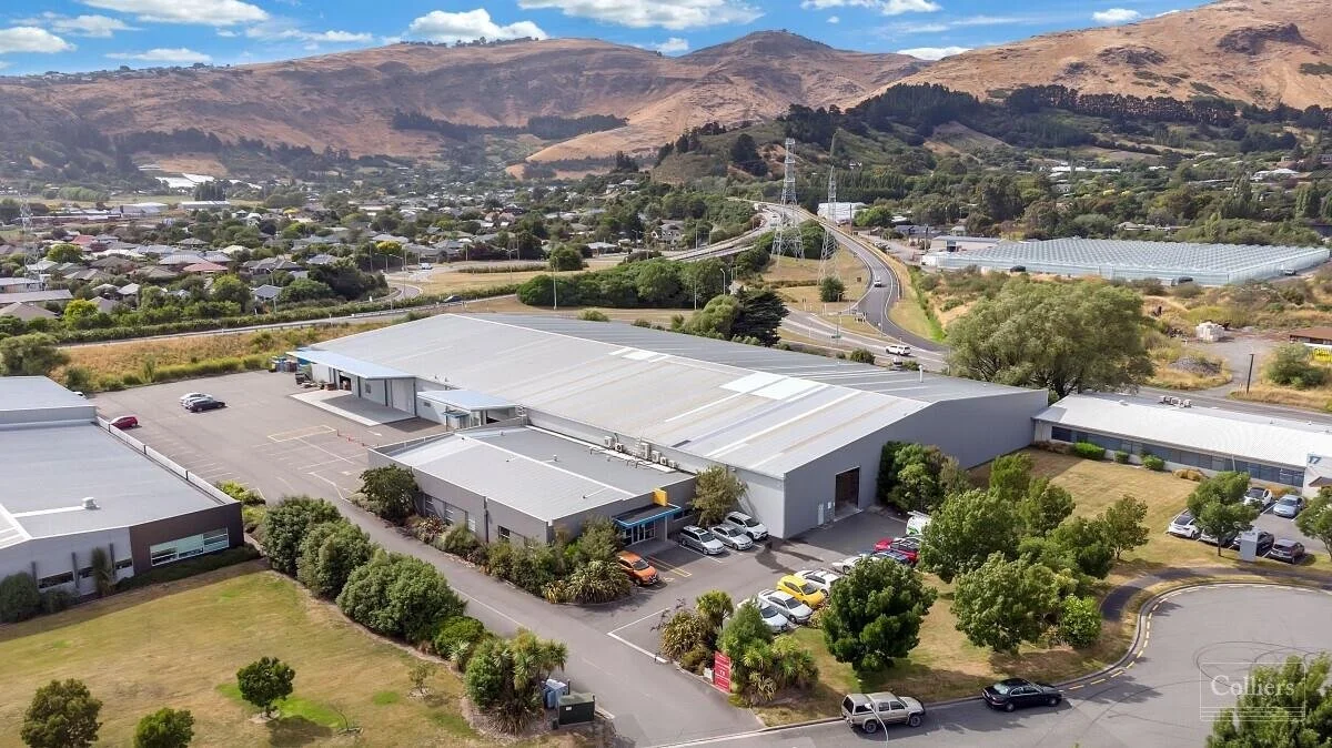 An aerial view of a commercial industrial complex with several buildings, parking lots, and surrounding green trees. In the background, there are hills and a road network connecting the area.